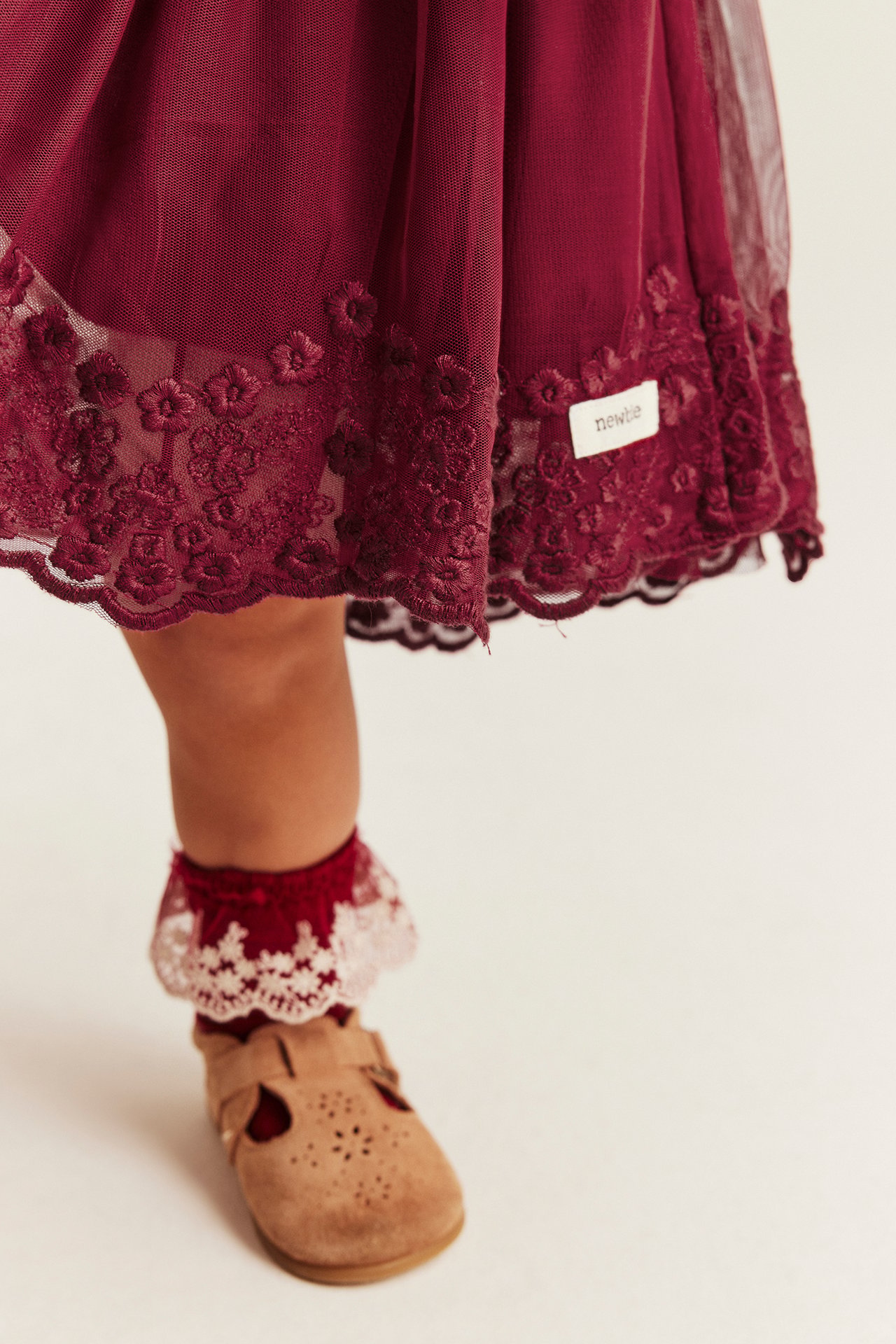 Close-up of a baby's maroon embroidered tulle dress, red lace-trim socks, and tan shoes.