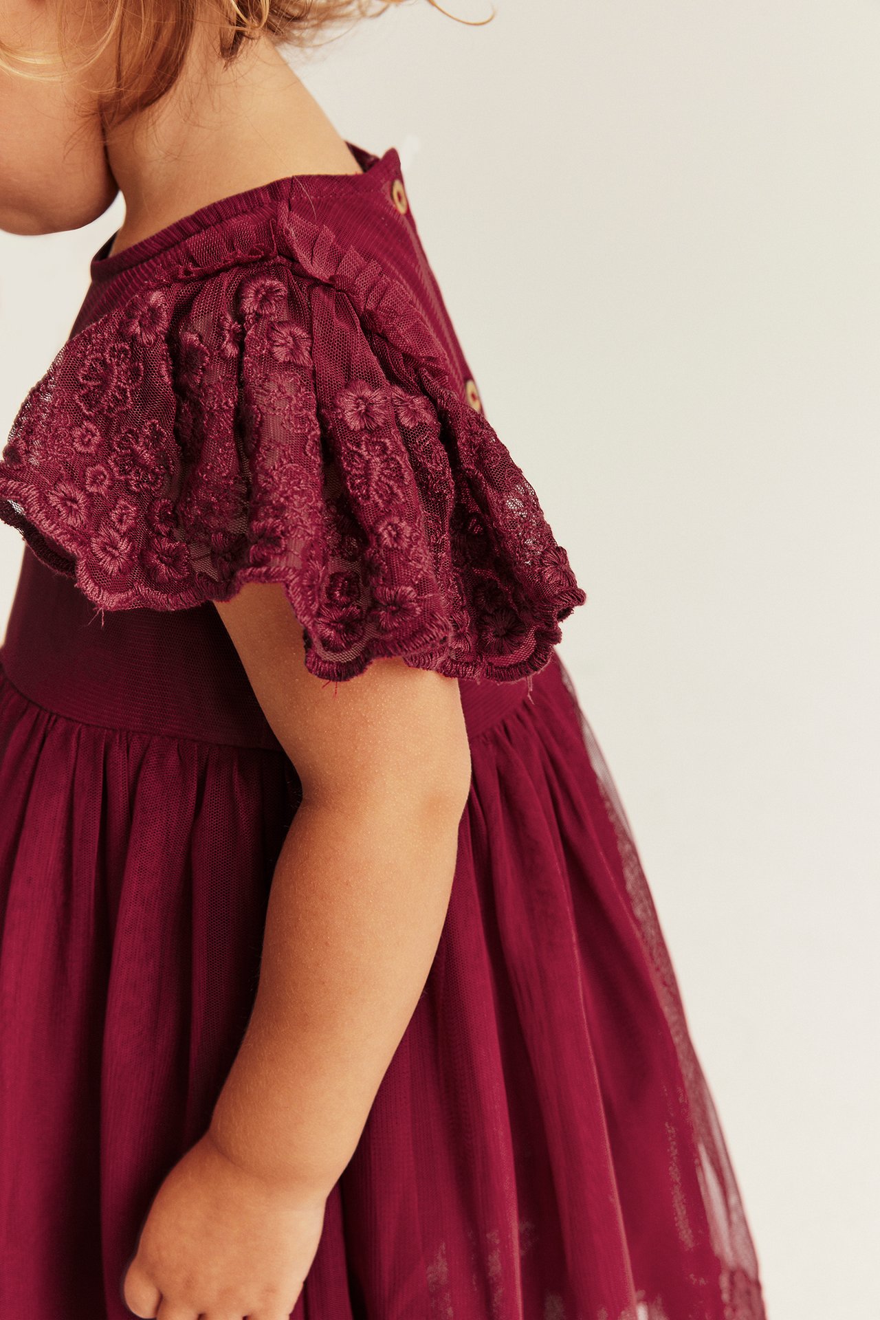 Close-up side view of a kid in a burgundy dress with ribbed top, lace sleeves, and tulle skirt.
