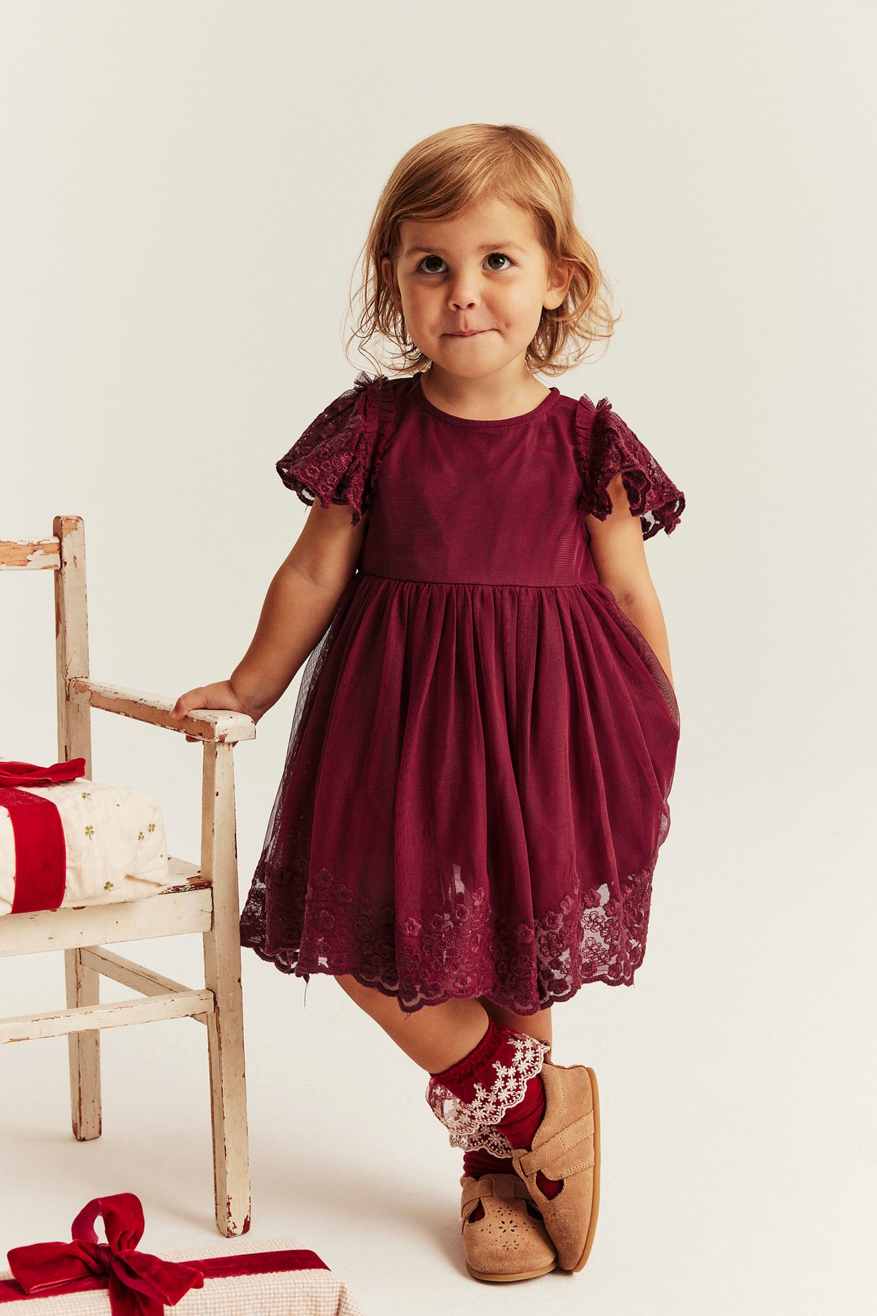 Vorderansicht Kind in bordeauxrotem Spitzenkleid, roten Spitzensocken und beigen Mary Jane Schuhen.