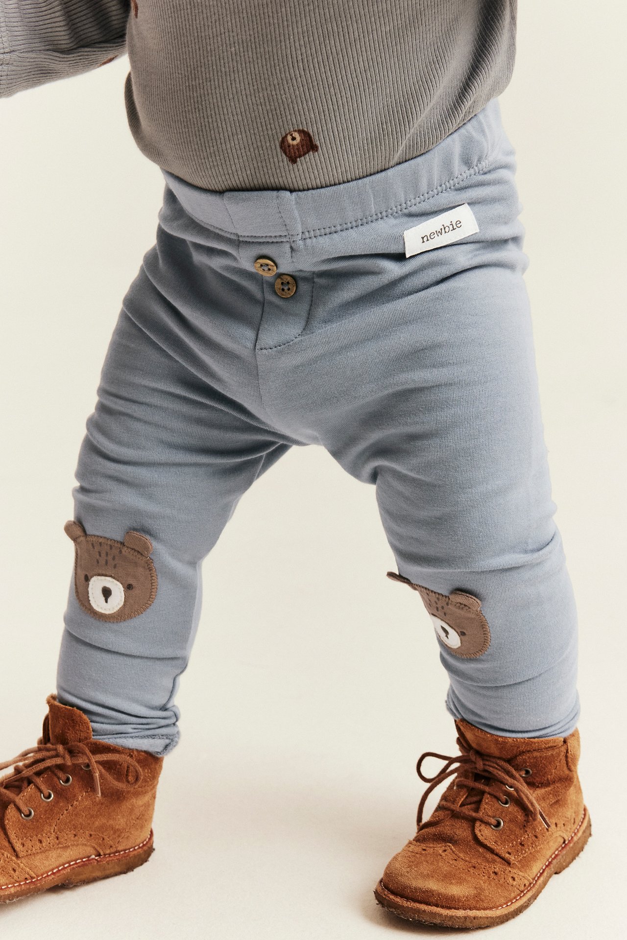 Vorderansicht eines Babys in blauer Hose mit Bärenknieflicken, grauem Oberteil und braunen Stiefeln.