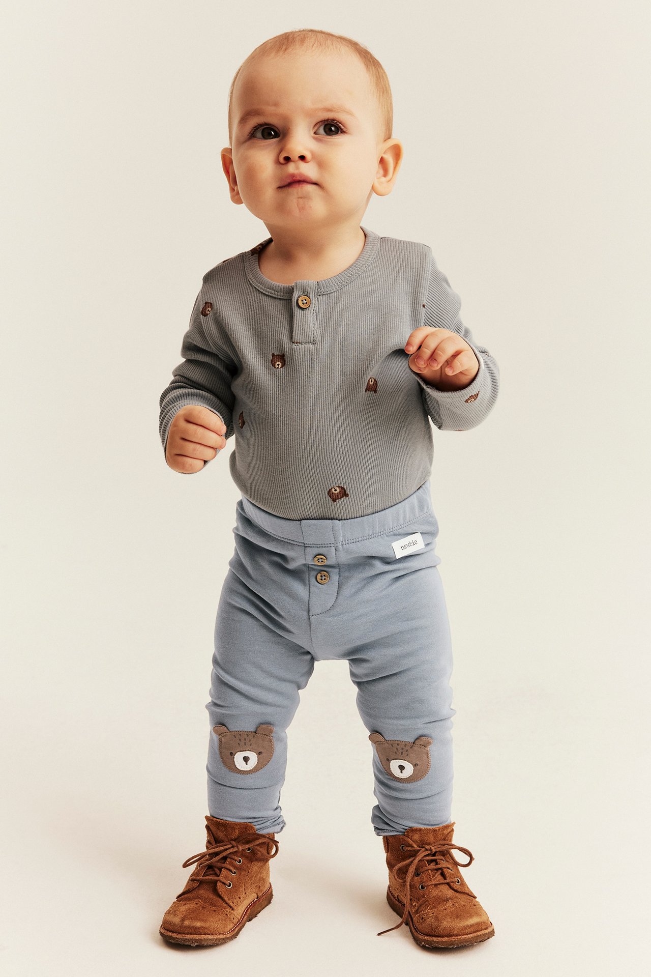 Baby in grauem besticktem Henley, hellblauen Leggings mit Bären-Knieflicken und braunen Schnürstiefeln, Vorderansicht.