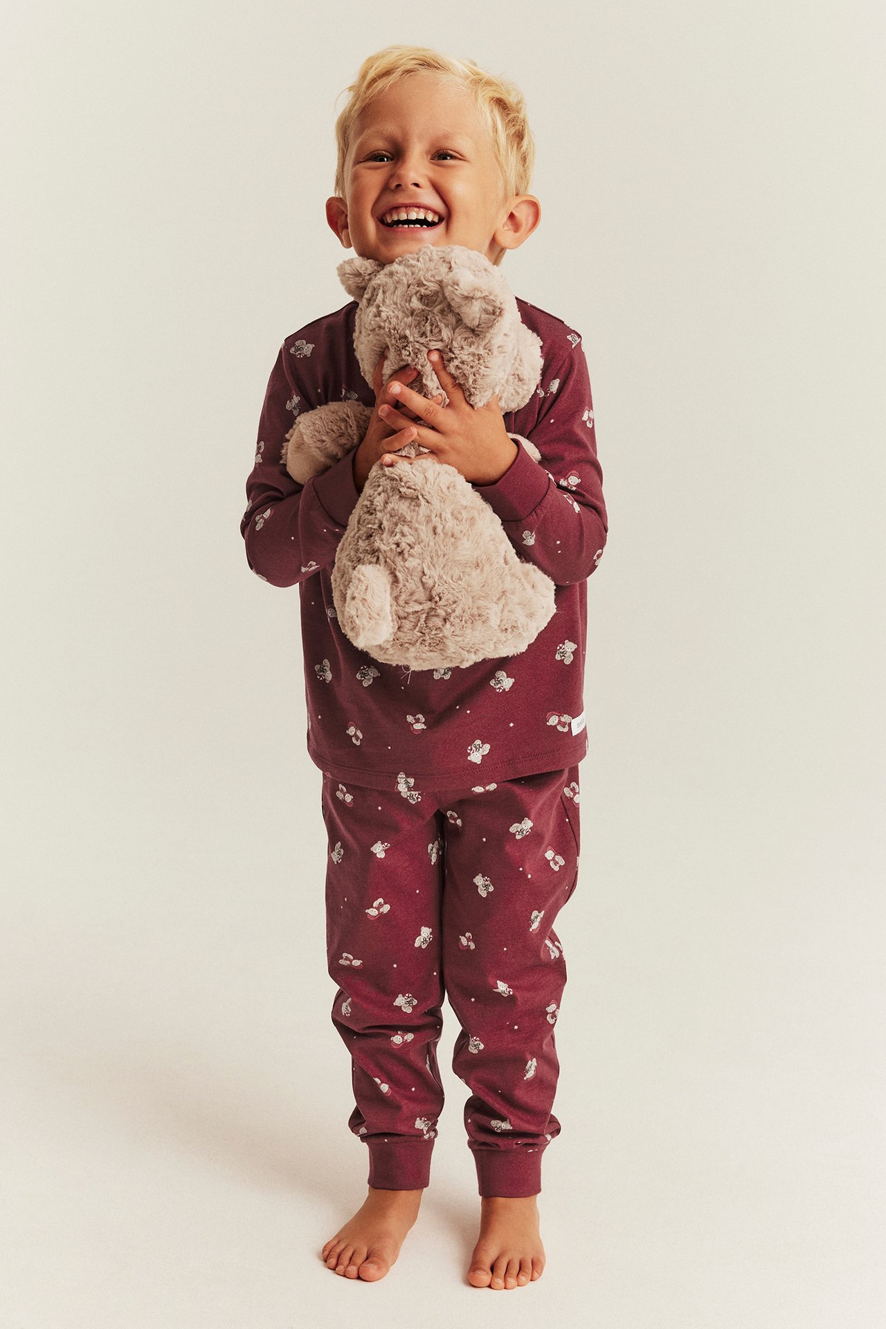 Frontansicht eines Kindes in burgunderrotem gemustertem Schlafanzug, hält einen hellbraunen Teddybären.