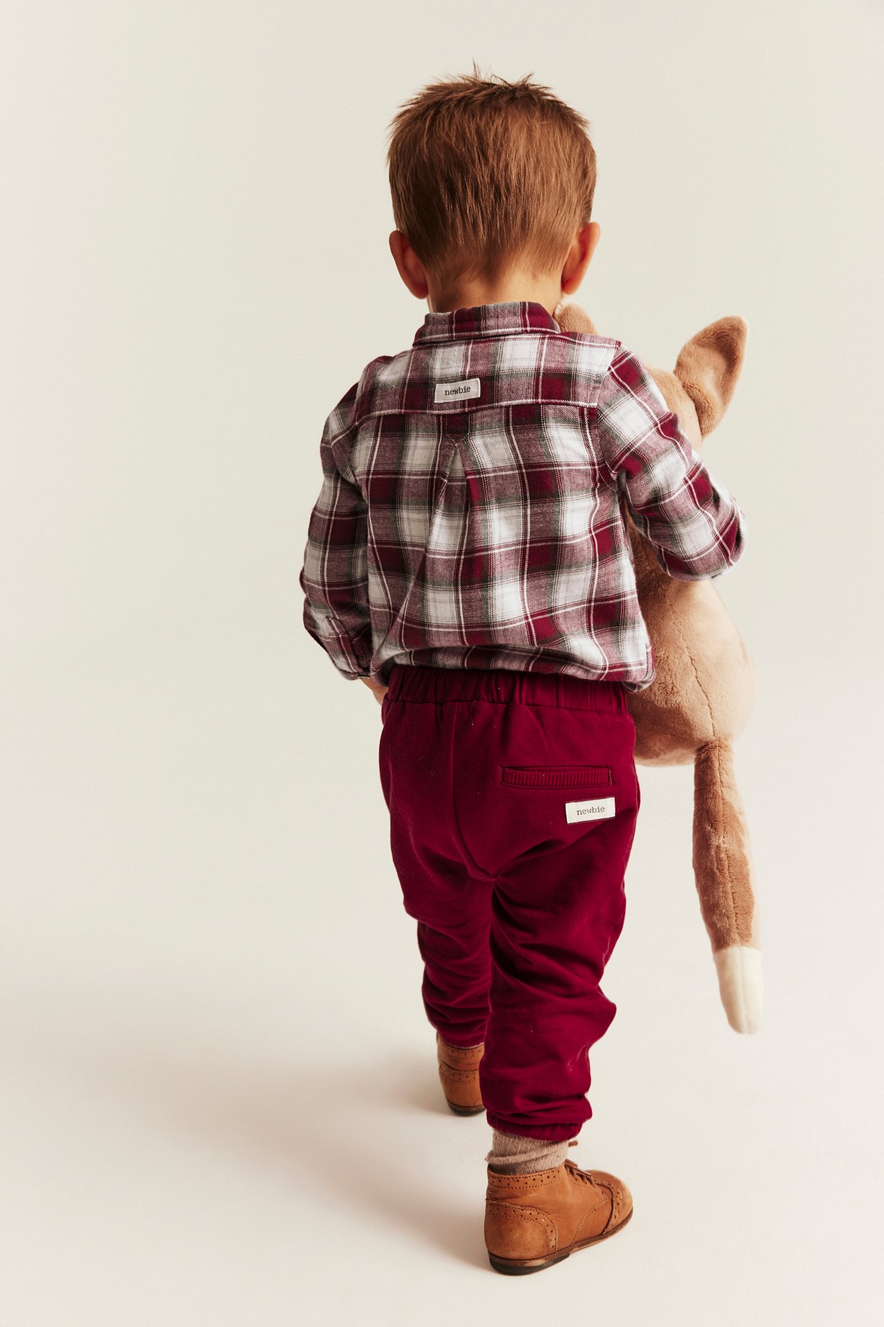 Back view of a kid in a red and white plaid shirt, red sweatpants, brown shoes, holding plush toy.