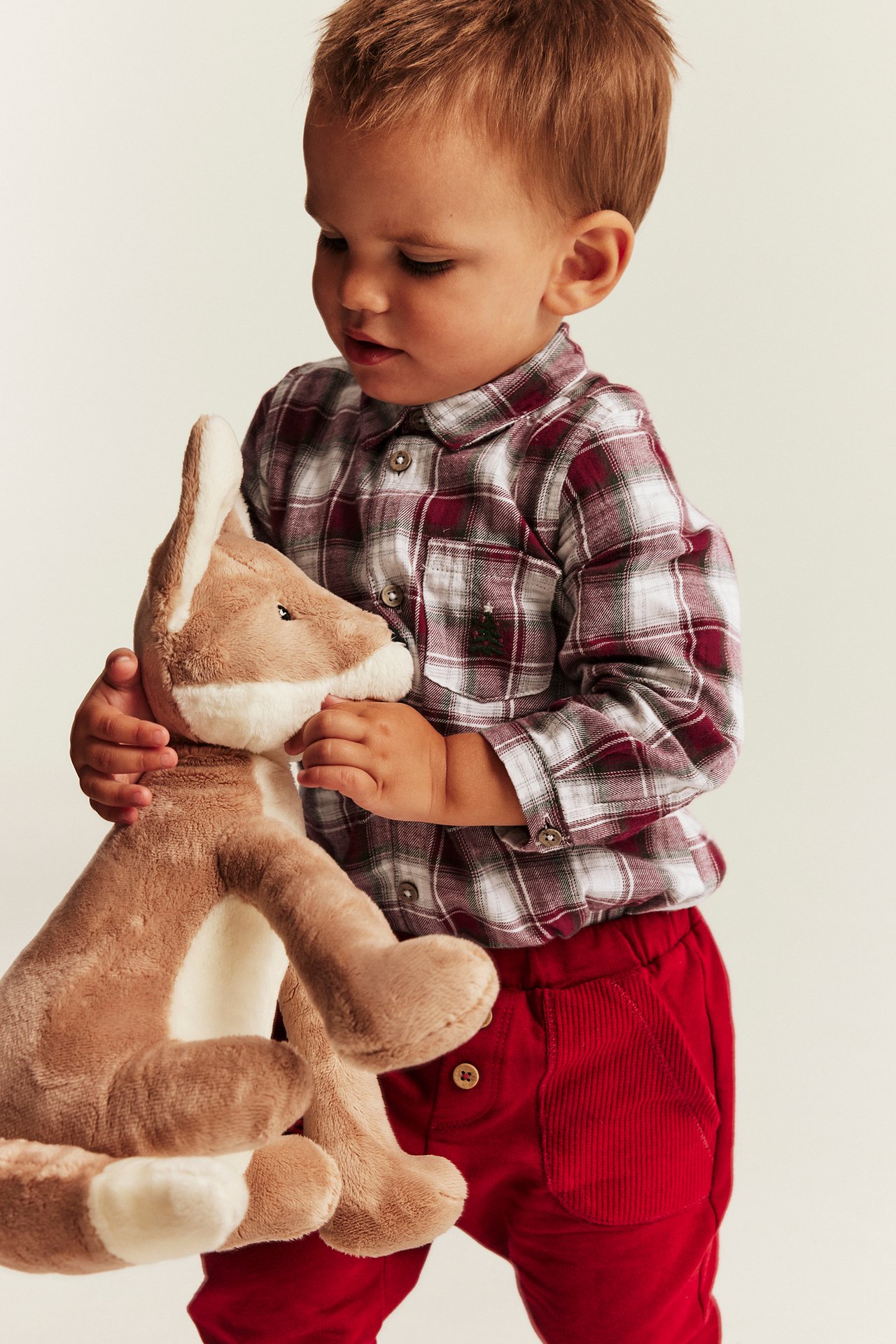 Baby in rot-weiß kariertem Langarmhemd, rote Cordhose, hält ein Stofftier.