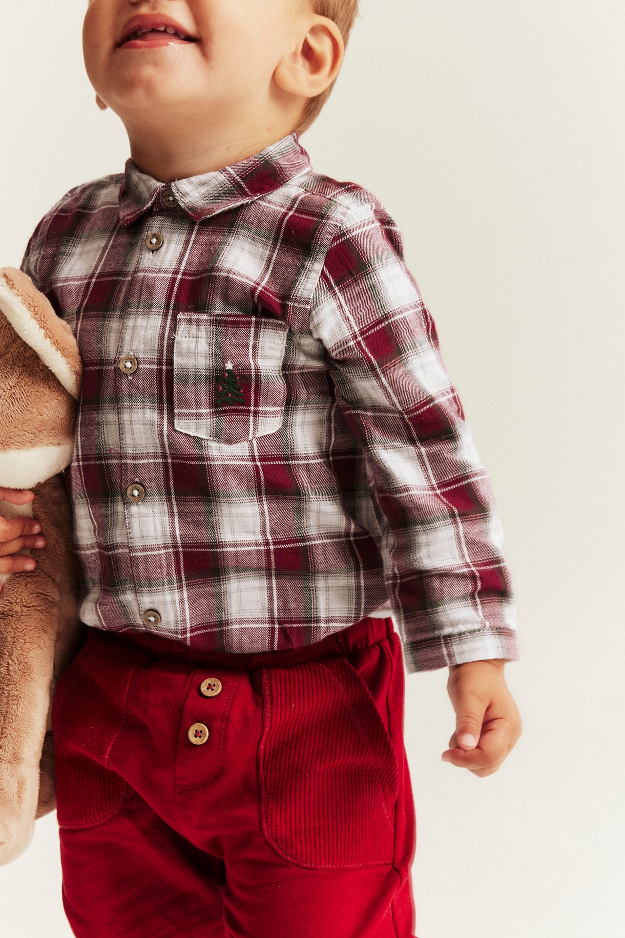 Nahaufnahme eines Babys in einem rot-weiß karierten Hemd mit Weihnachtsstickerei und roter Cordhose.