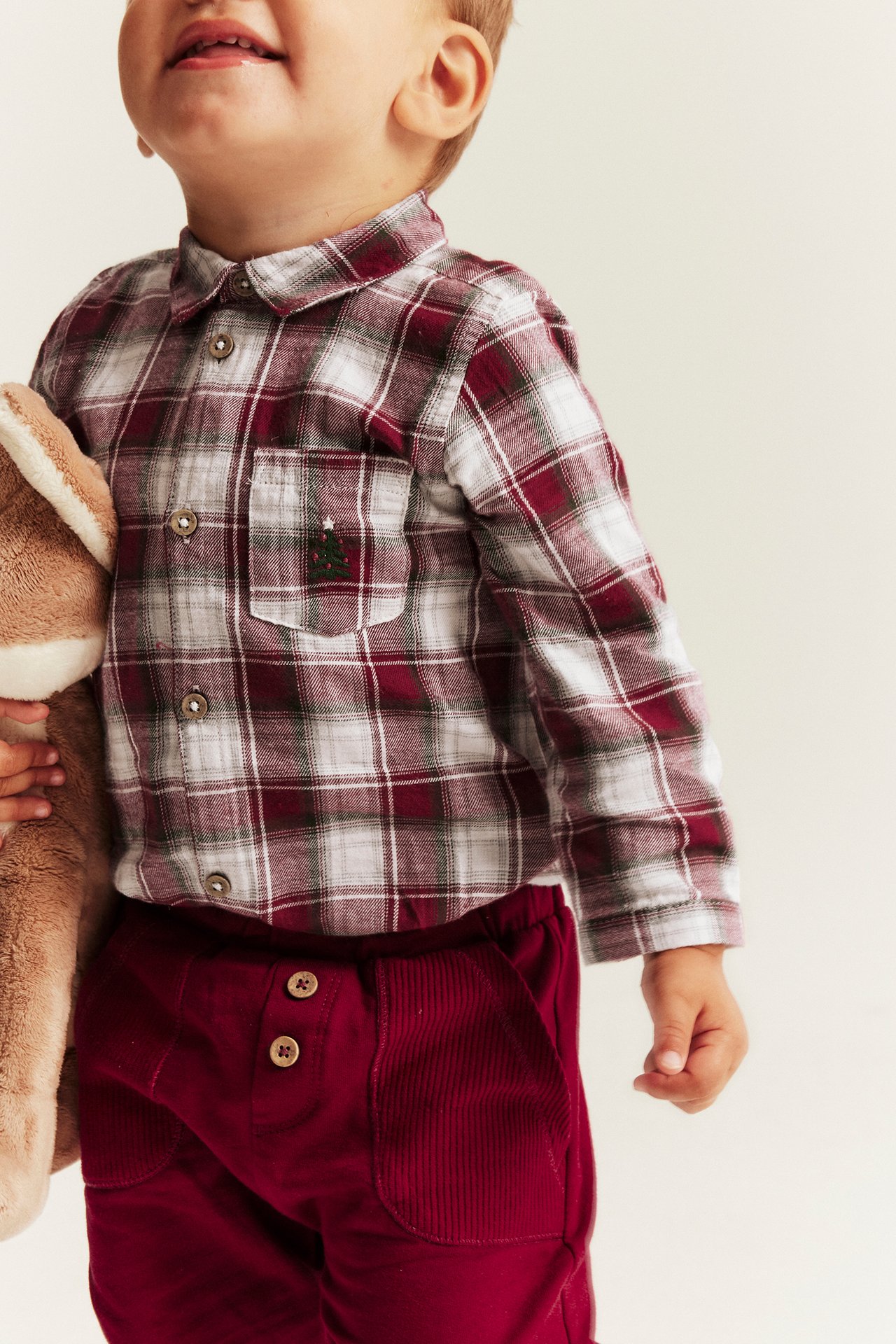 Front view: Kid in red and white plaid shirt with tree embroidery, red corduroy pants, holding a toy.
