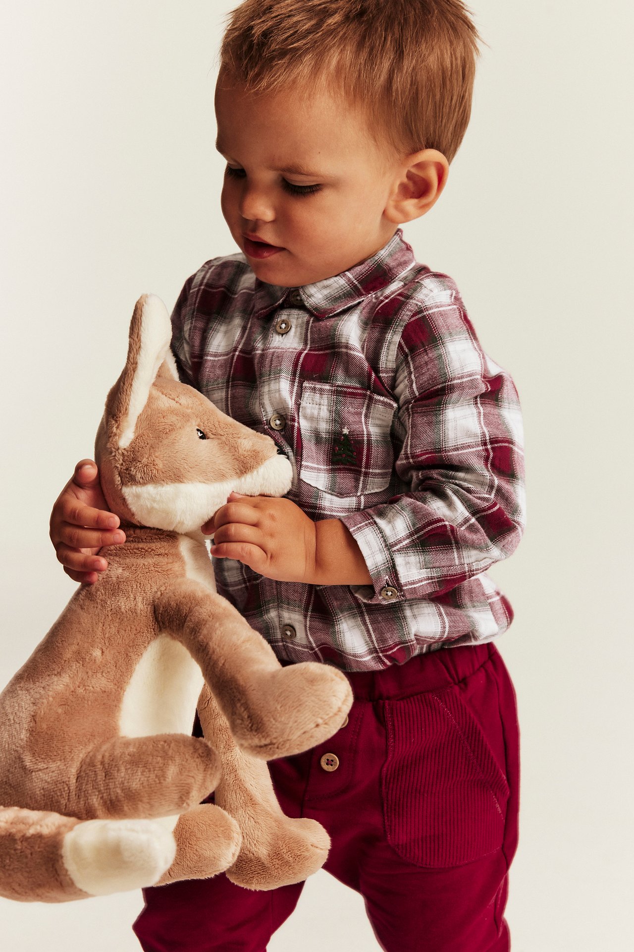 Front view of a baby in a red plaid shirt and red corduroy pants, holding a plush fox.