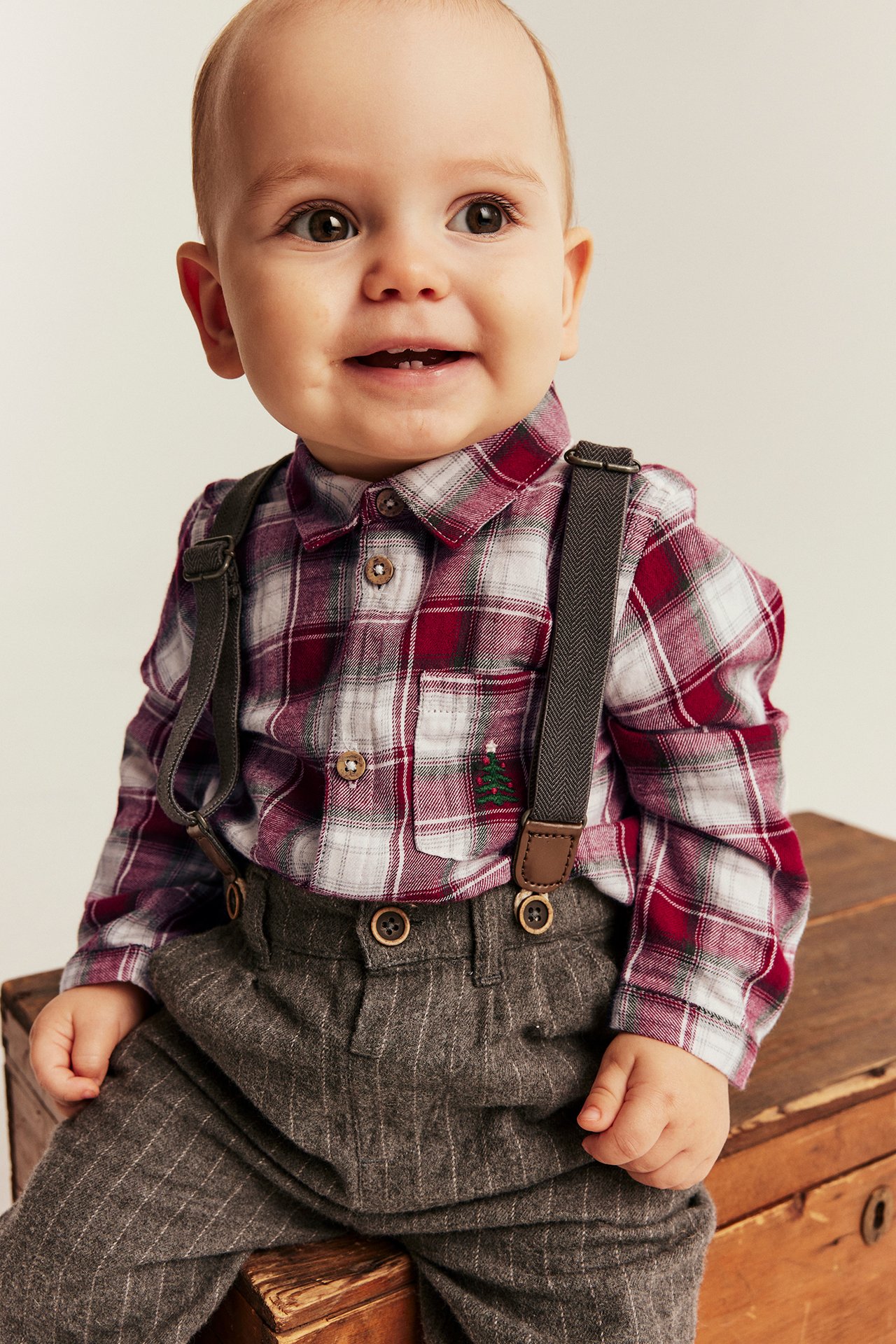 Front view, baby boy in red plaid shirt with Christmas tree, gray suspenders, and pinstripe trousers.