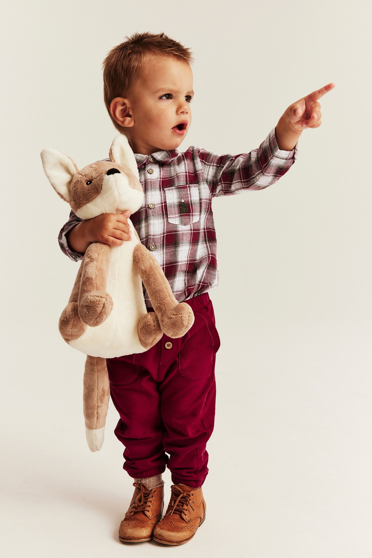Front view: Kid in red and white plaid shirt, red corduroy pants, brown lace-up boots, holding plush fox.