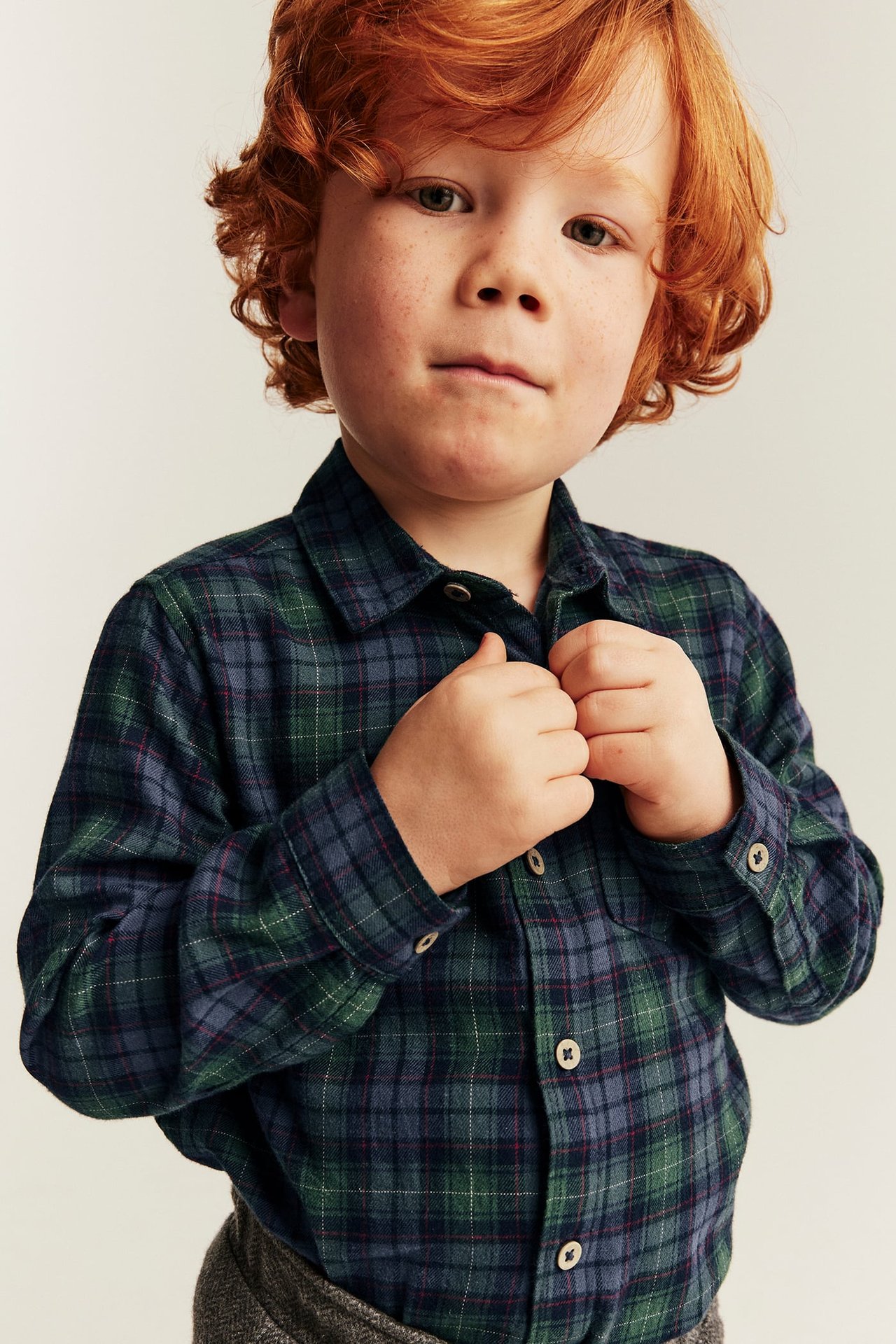 Front view of a kid adjusting a dark green and blue plaid flannel shirt over grey patterned pants.