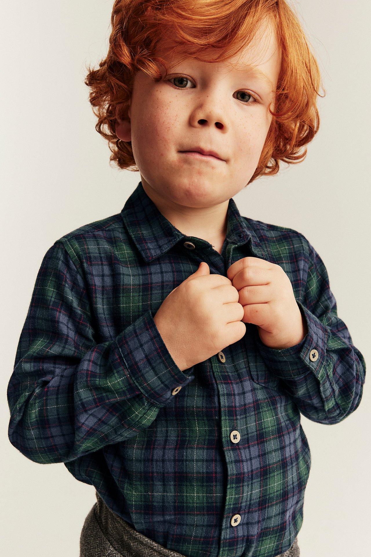 Front view of a kid adjusting a dark green and blue plaid flannel shirt over grey patterned pants.
