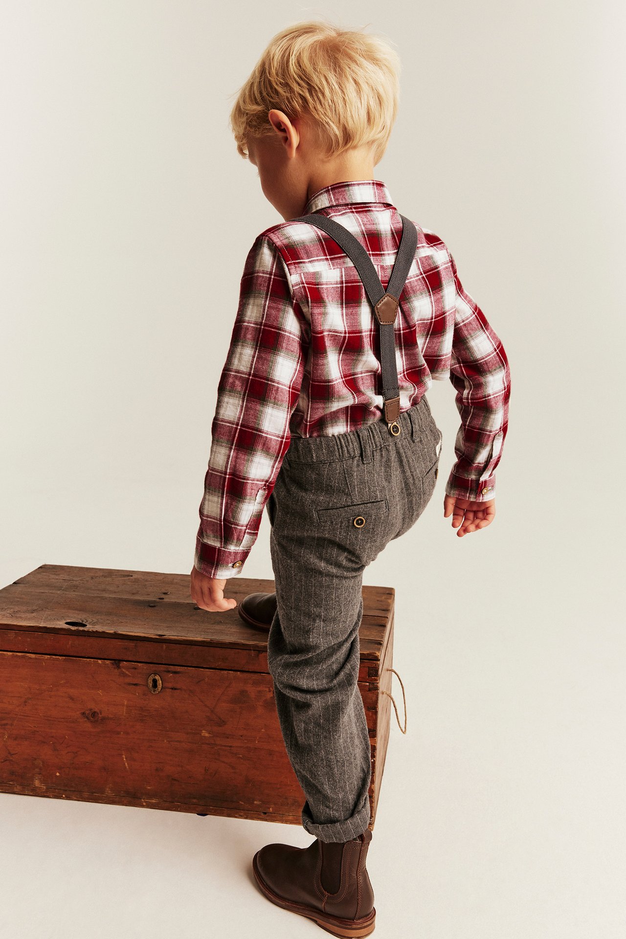 Back view: kid wearing red plaid shirt, grey pinstripe trousers with suspenders, brown boots.