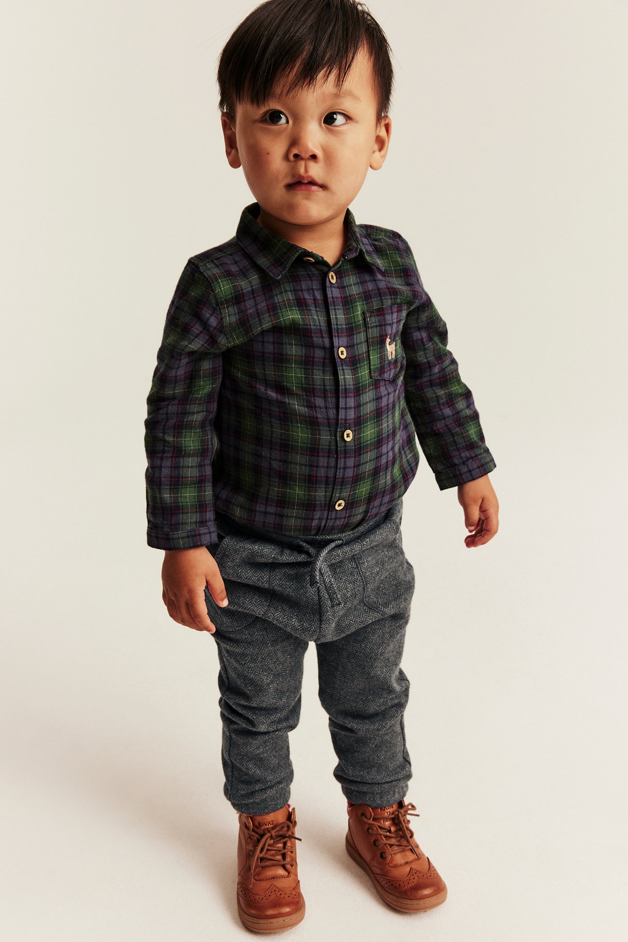 Front view of a boy wearing a plaid button-down shirt, gray drawstring trousers, and brown lace-up boots.