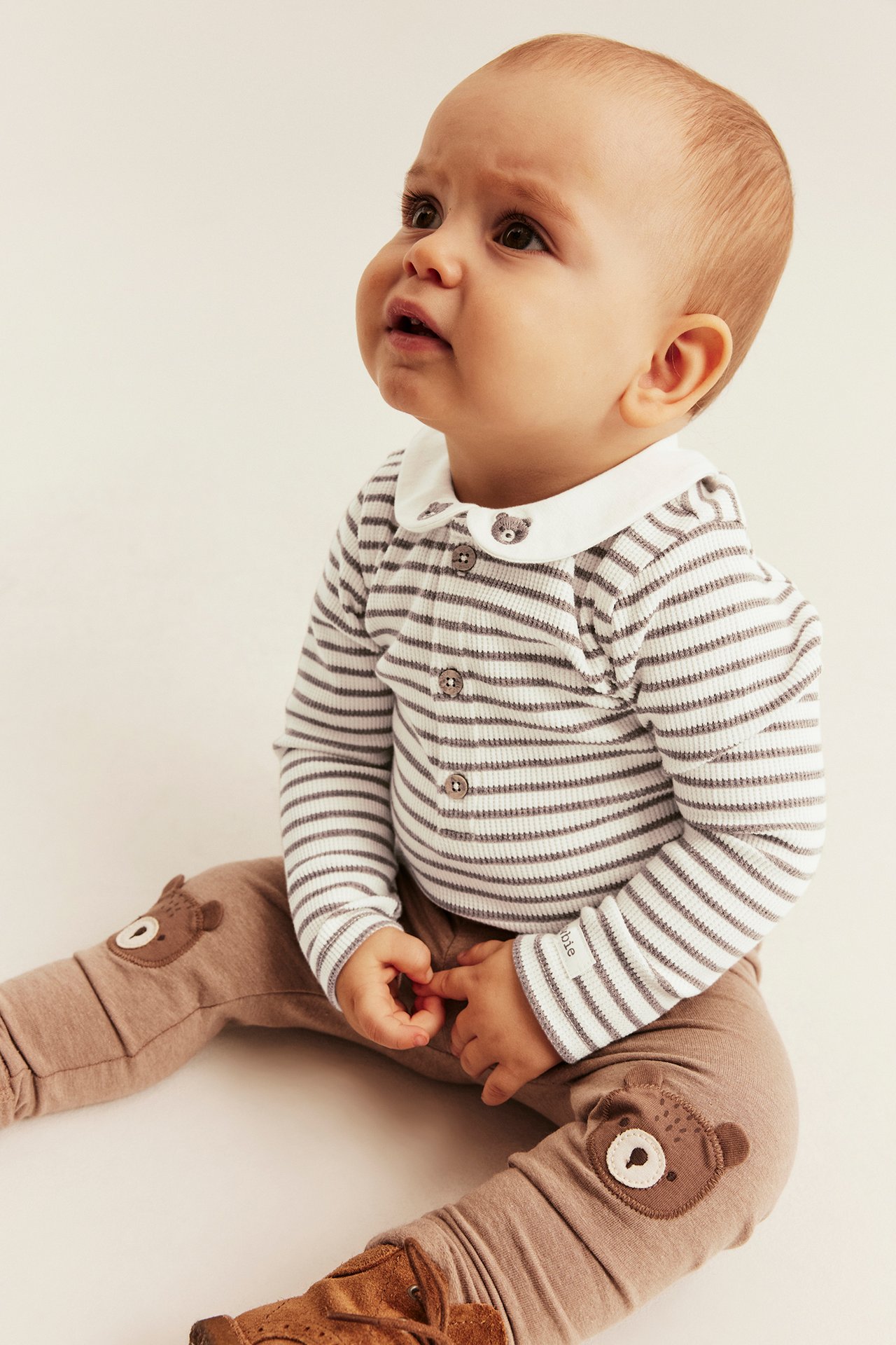 Front view of a baby in a ribbed striped top, brown pants with bear patches, and brown booties.