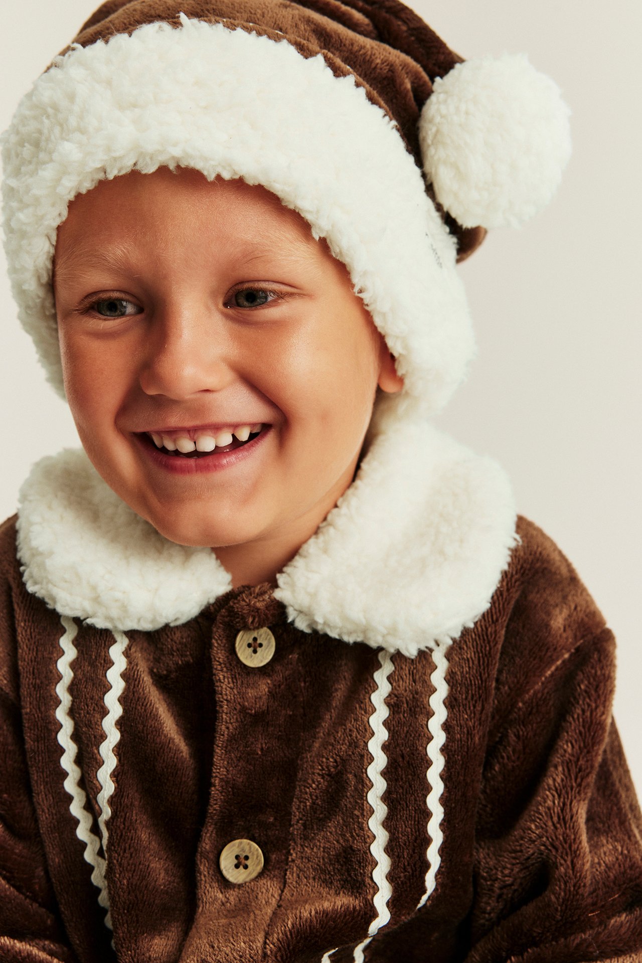 Nahaufnahme frontal eines Kindes mit brauner Plüsch-Lebkuchen-Weihnachtsmütze und passendem Oberteil.