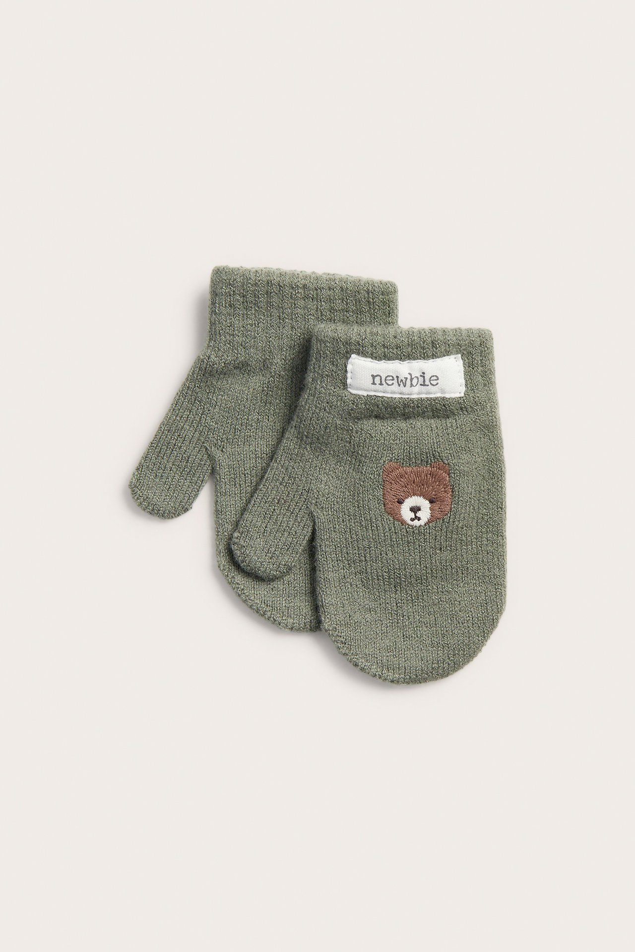 Draufsicht auf ein Paar grüne gestrickte Babyfäustlinge, einer mit brauner Bärenstickerei.