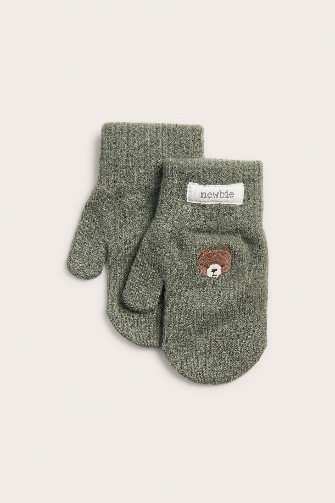 Vorderansicht olivgrüner gestrickter Babyfäustlinge mit kleiner brauner Bärenstickerei und Newbie-Etikett.