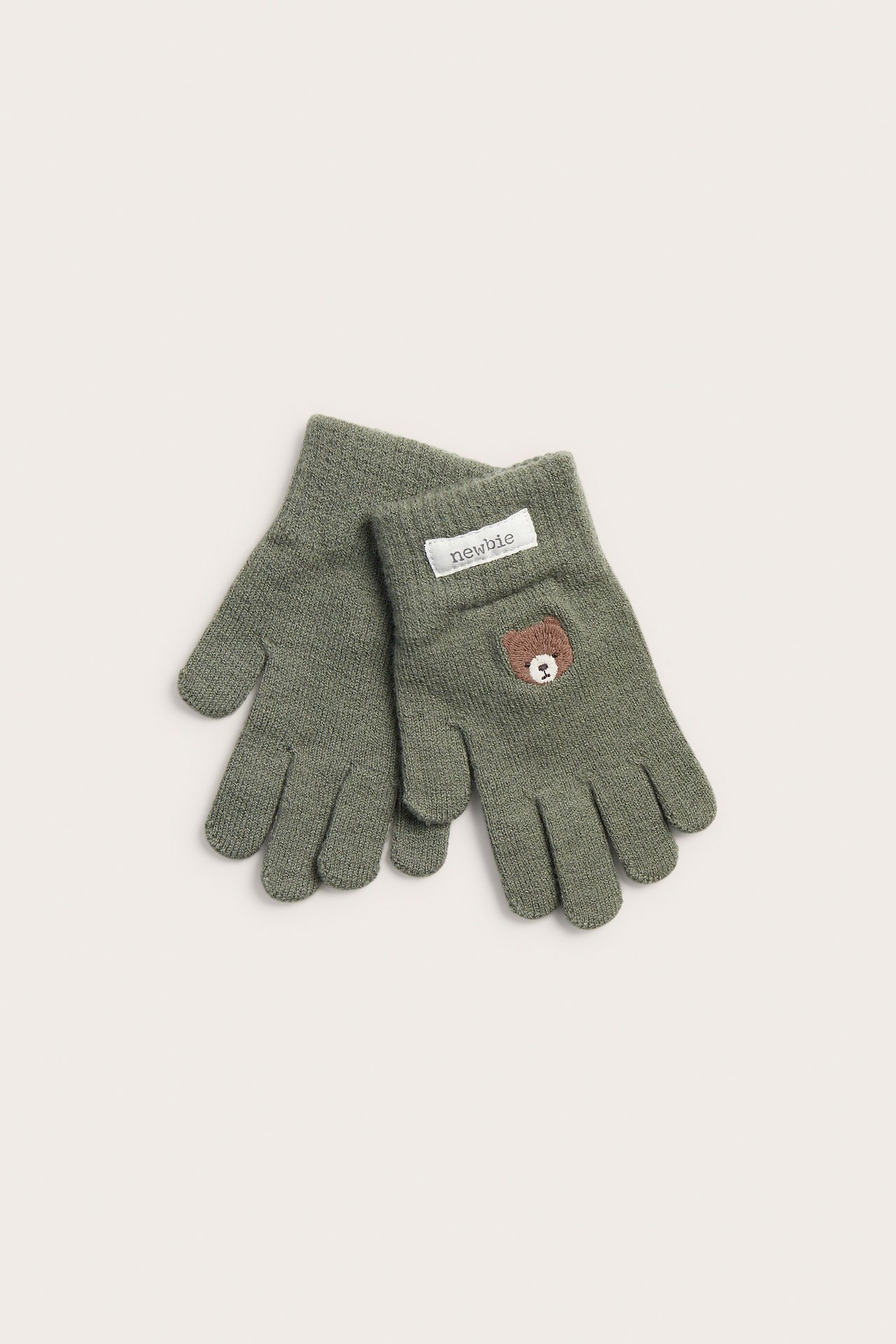 Draufsicht auf ein Paar grüne gestrickte Kinderhandschuhe, einer mit Newbie-Etikett und Bärenstickerei.
