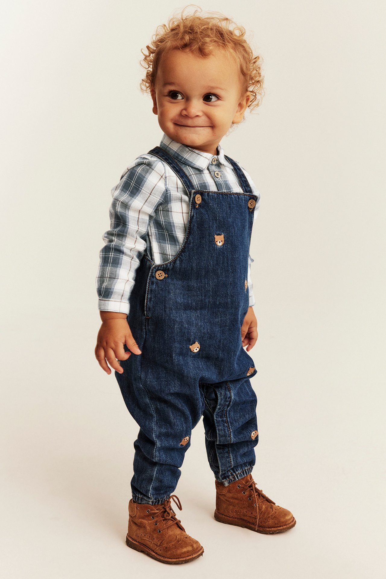 Front view, baby in dark blue denim overalls with bear embroidery, plaid shirt, and brown suede boots.