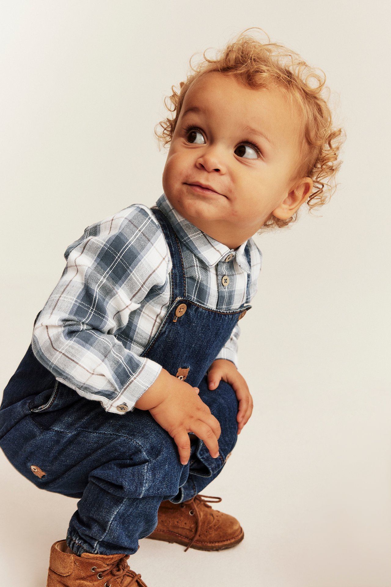 Front view of a baby wearing dark blue denim overalls, a blue and white plaid shirt, and brown suede boots.