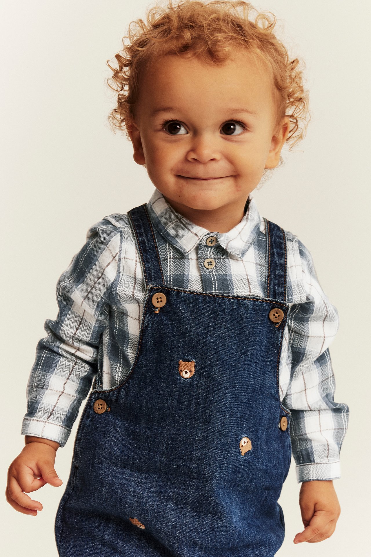 Front view of a baby wearing dark blue denim dungarees with bear embroidery over a plaid shirt.