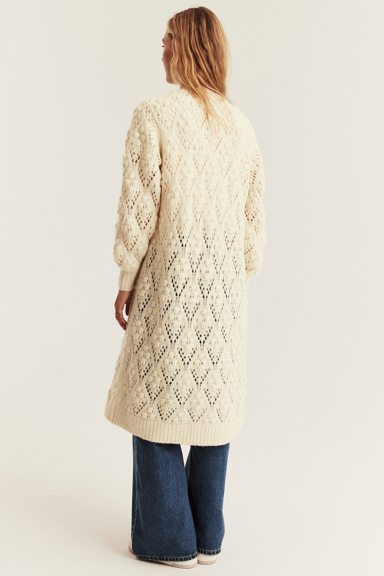 Back view of a woman wearing a long off-white textured knit cardigan, dark blue wide-leg jeans, and white sneakers.