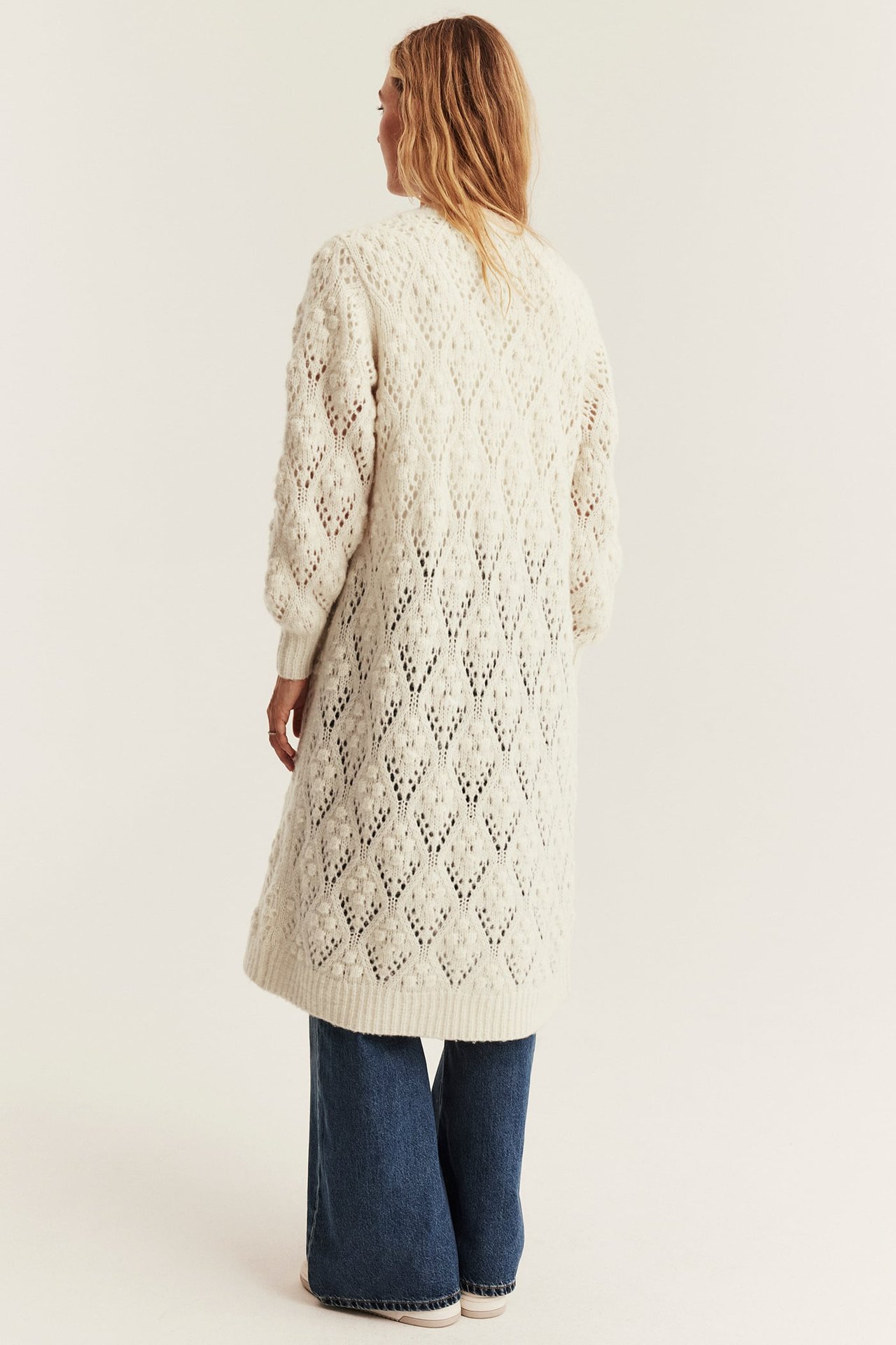 Back view of a woman in a long white knitted cardigan with open-knit pattern, paired with blue jeans.