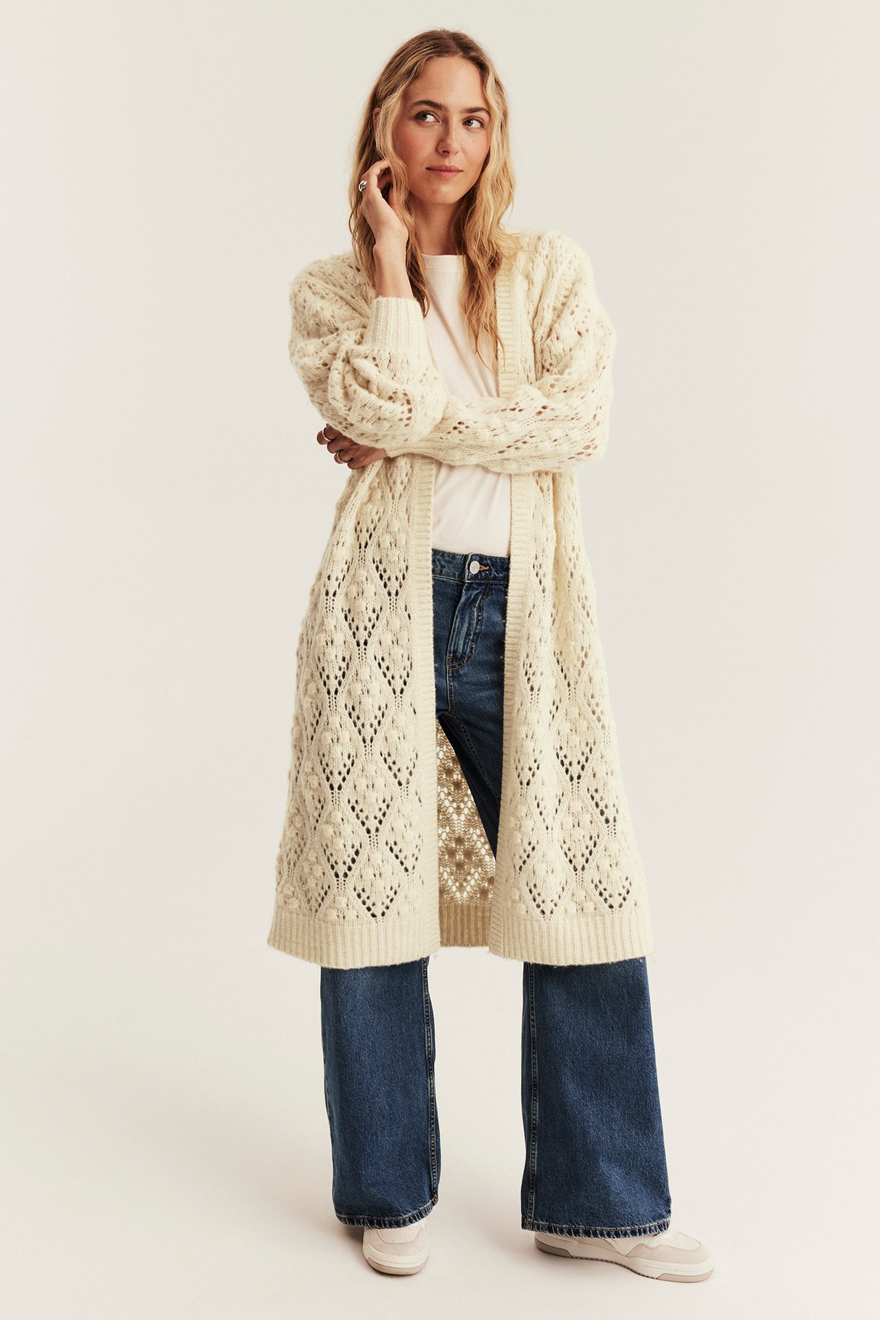 Front view of a woman wearing a long cream patterned knit cardigan, white t-shirt, blue jeans, and sneakers.