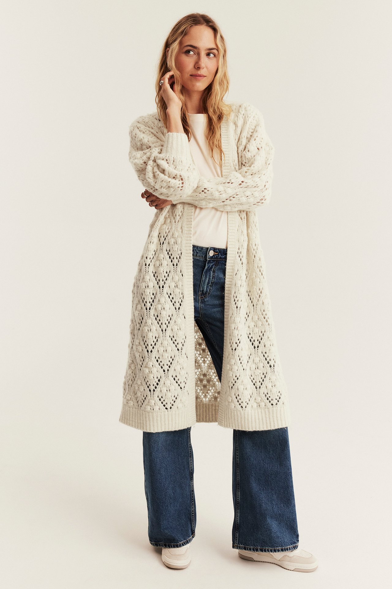 Vorderansicht einer Frau in langem cremefarbenem Strickcardigan über weißem T-Shirt und blauer Jeans.