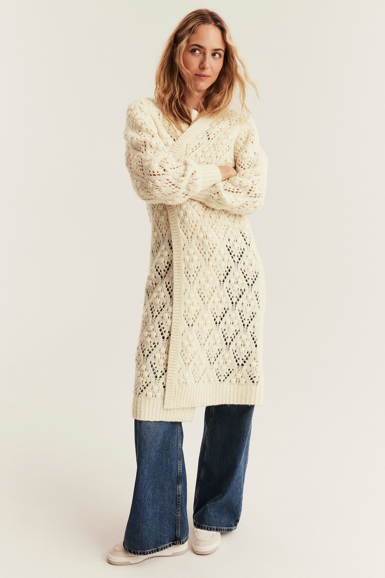 Front view of a female model wearing a long cream knitted openwork cardigan, blue jeans, and white sneakers.