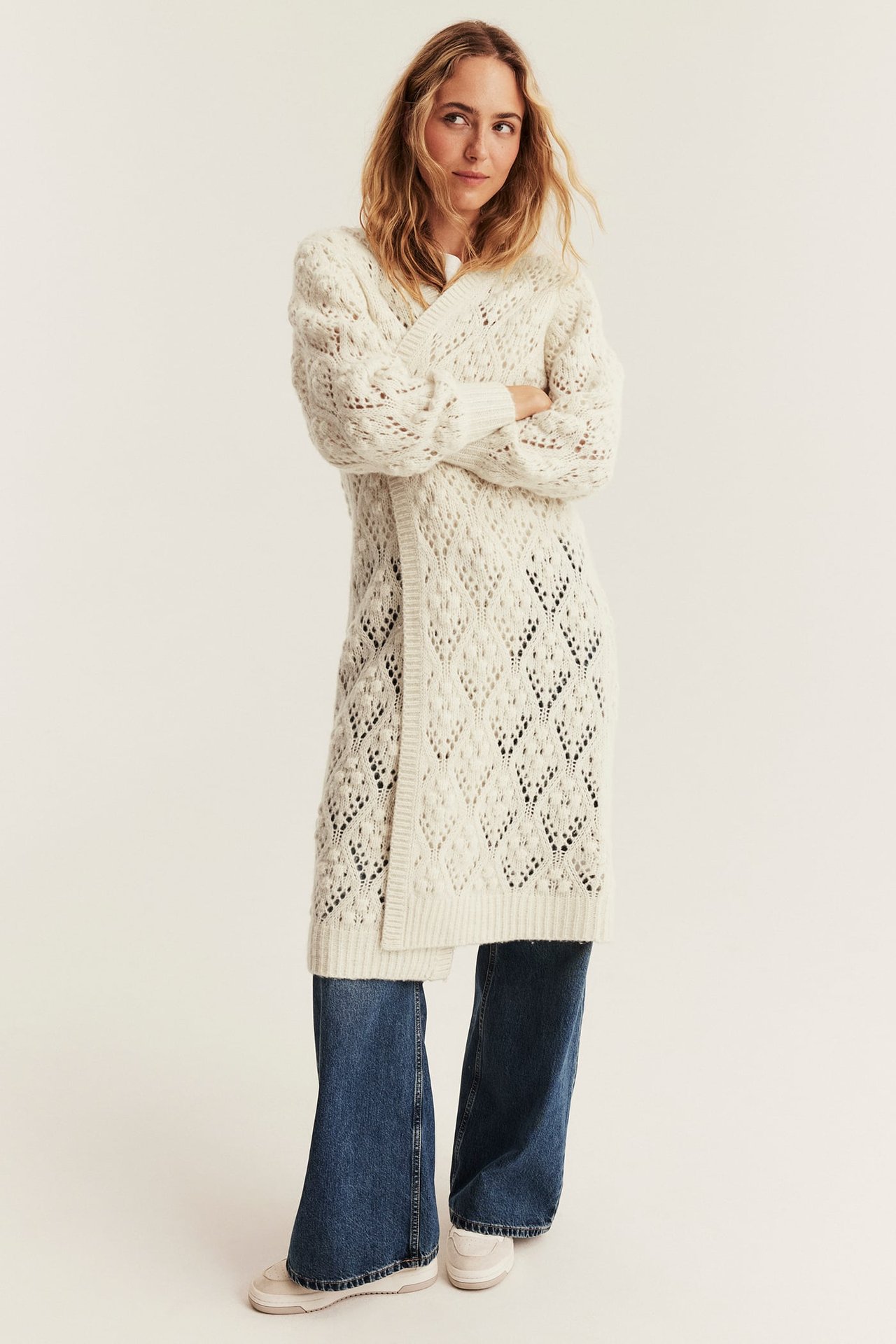 Female model in a long cream openwork knit cardigan, blue wide-leg jeans, and light sneakers, front view.