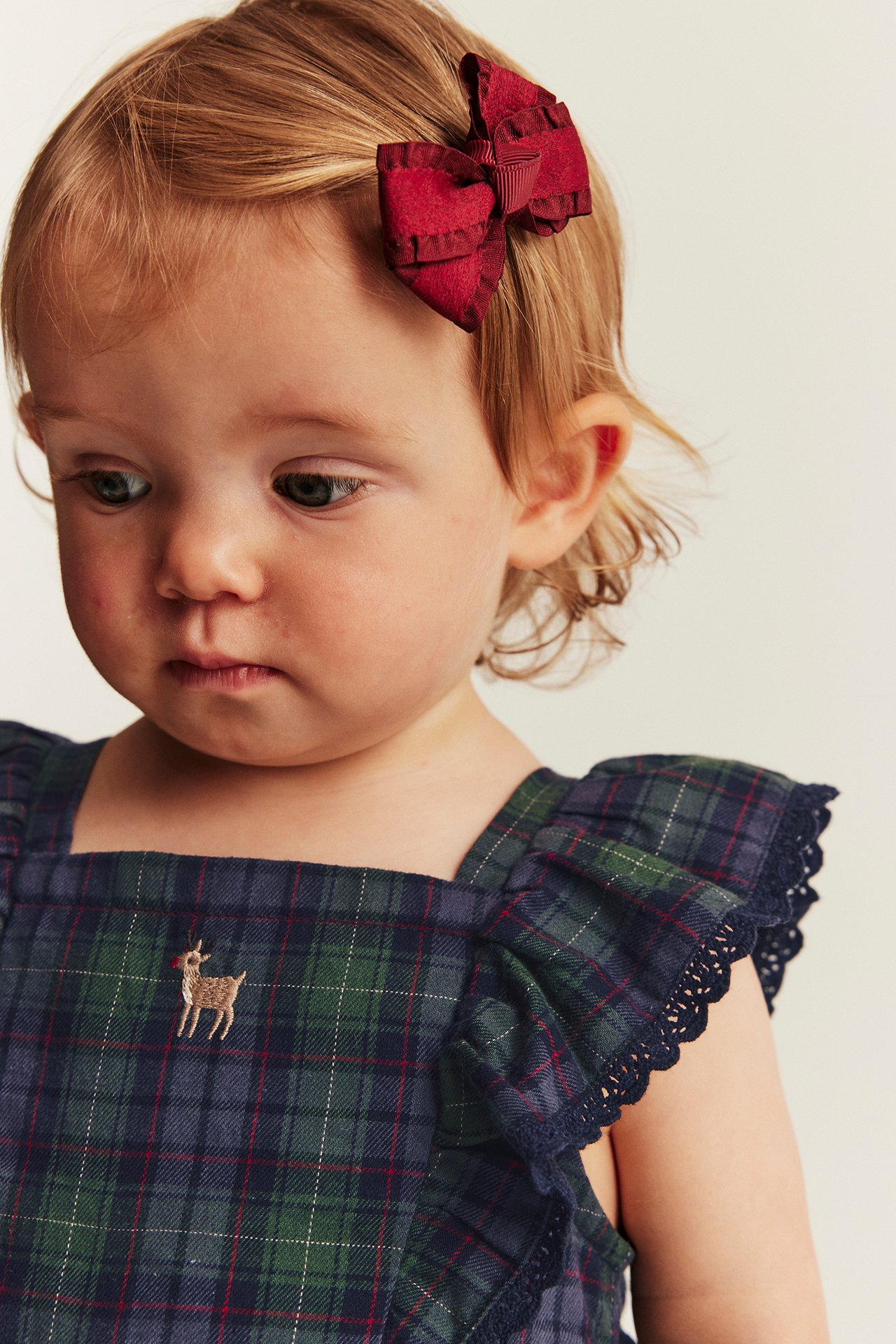 Front view of a baby in a blue-green tartan romper with embroidered deer and red hair bow.