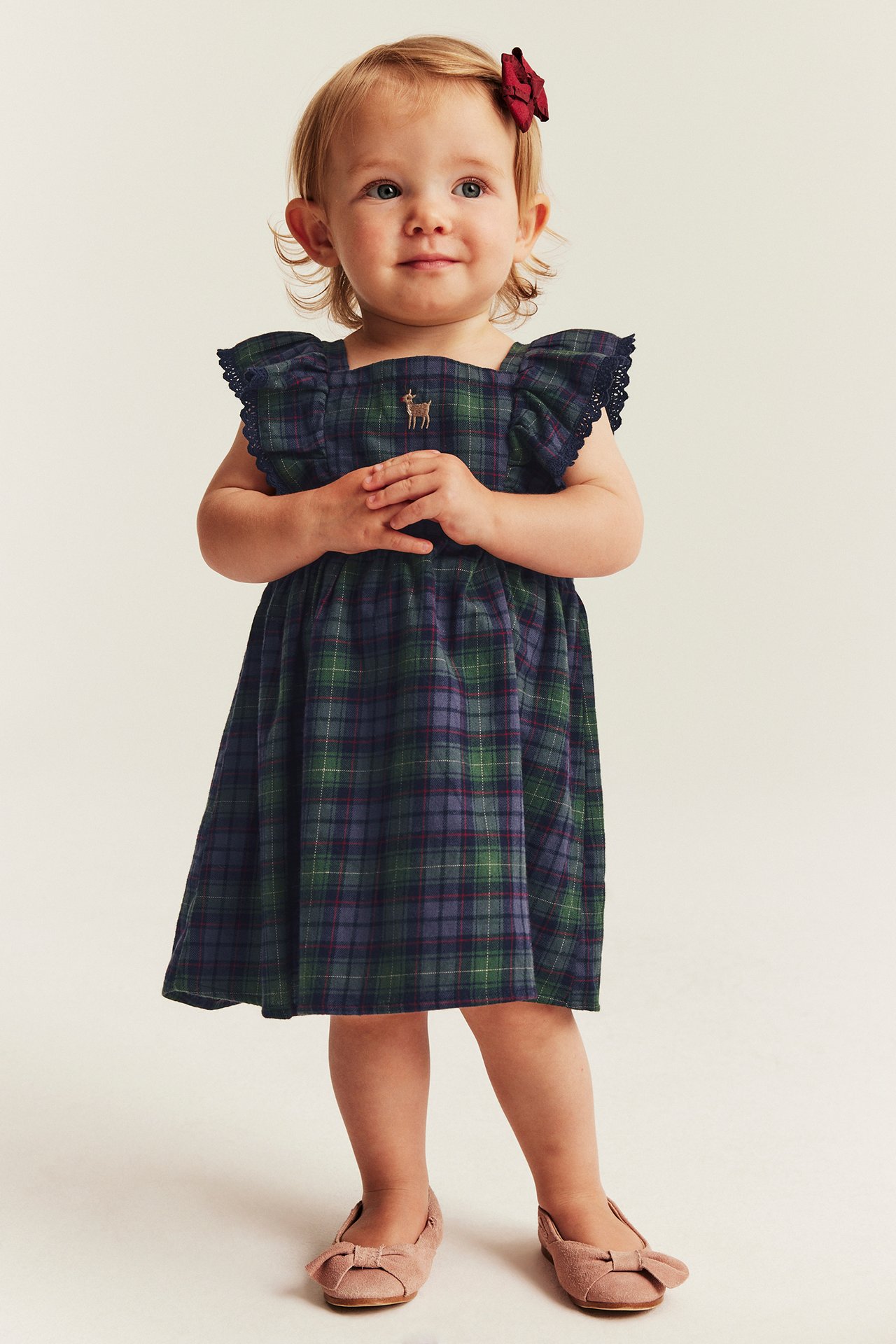 Front view of a baby girl wearing a green plaid dress with deer embroidery and pink bow shoes.