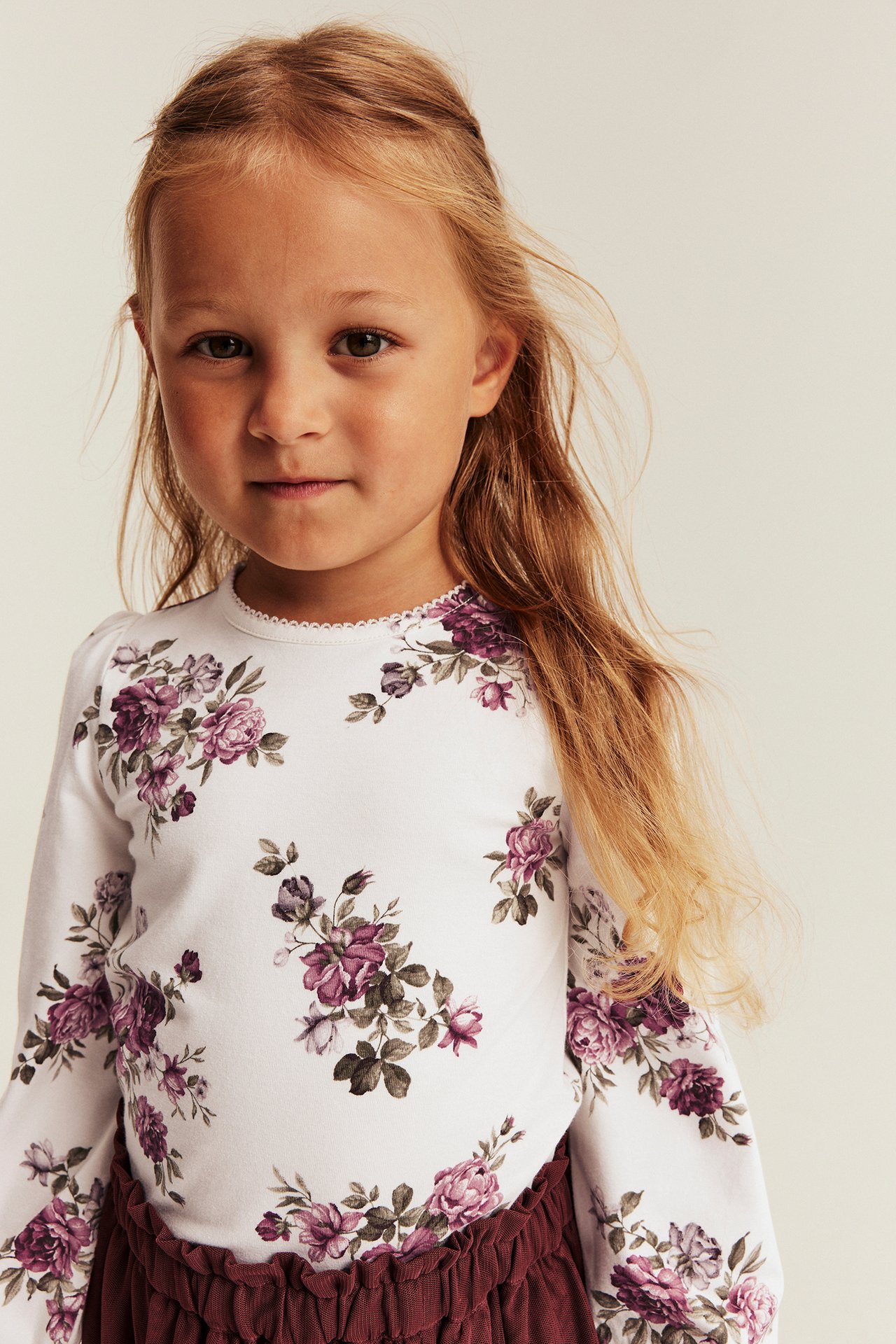 Front view of a kid wearing a white long-sleeved top with purple floral print and a burgundy ruffled skirt.
