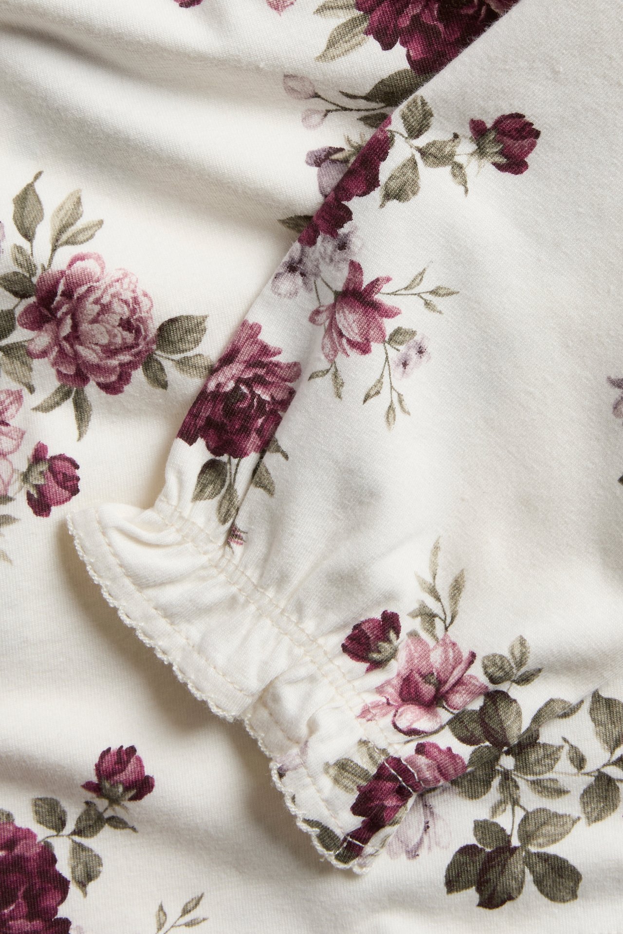 Close-up of a cream floral print fabric with ruffled trim featuring dark red roses and green leaves.