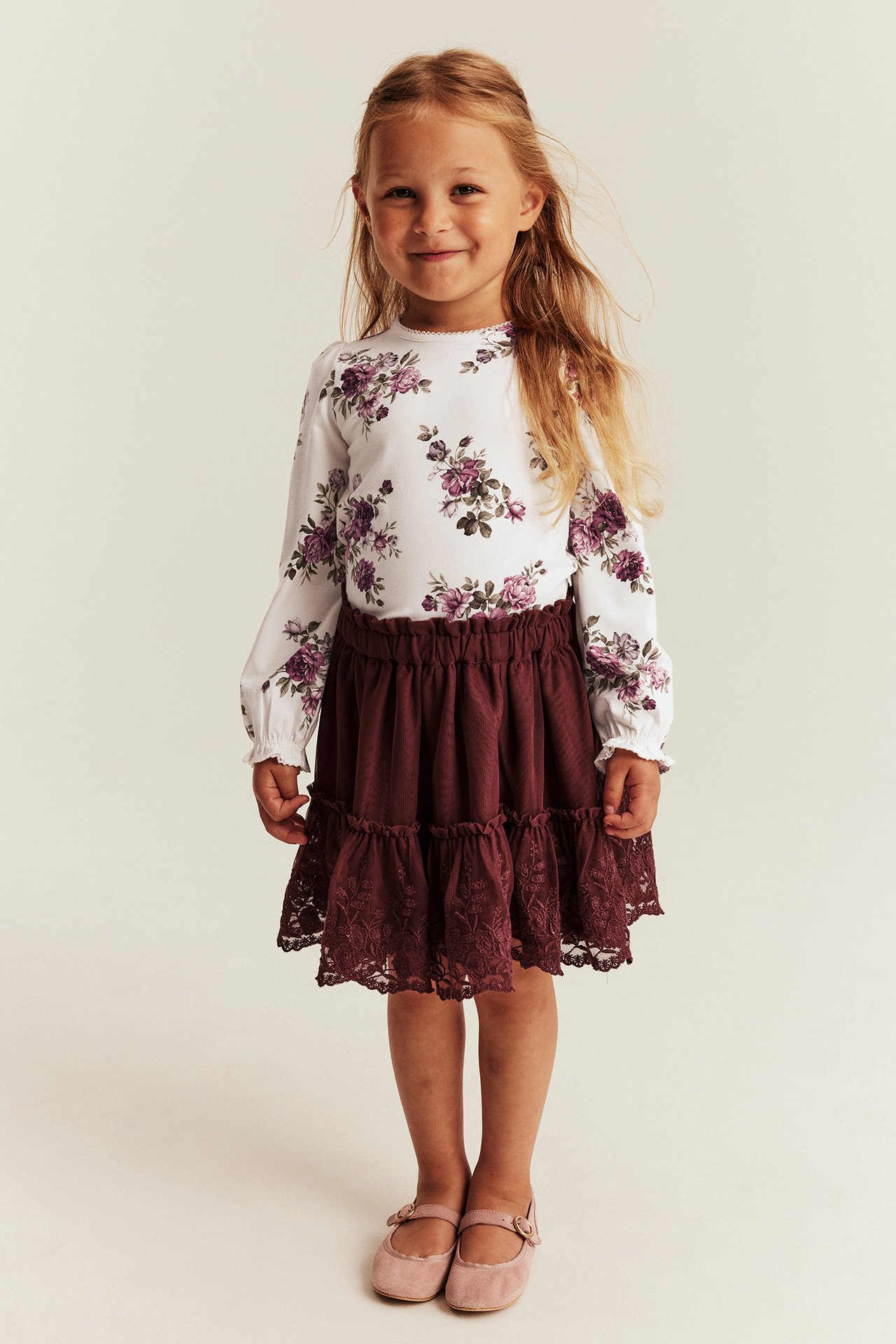 Vorderansicht: Kind trägt einen burgunderfarbenen Stufenrock mit Spitze, ein weißes Blumen-Langarmshirt und rosa Schuhe.