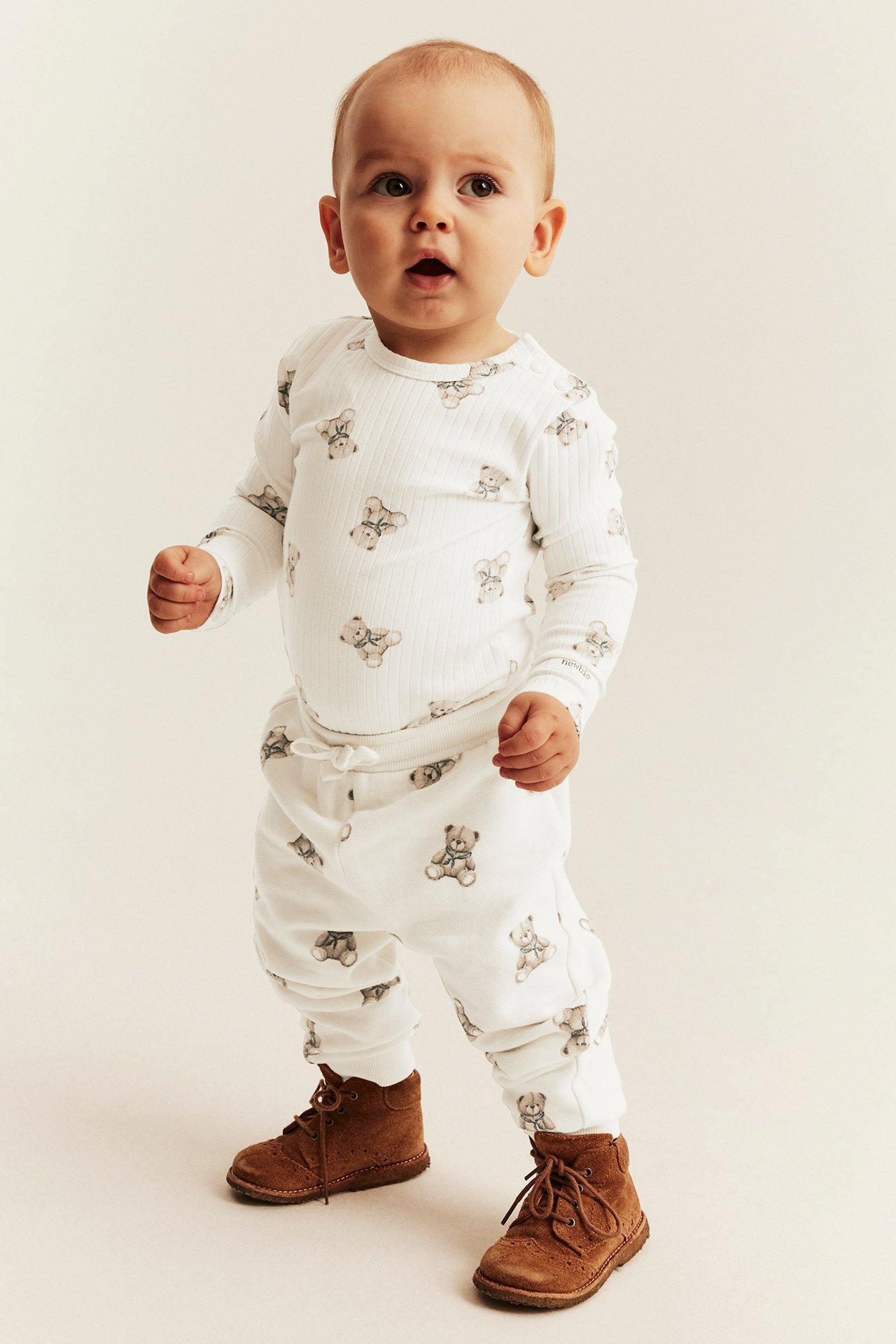 Front view of baby wearing a white ribbed teddy bear print long-sleeve bodysuit, matching pants, and brown boots.