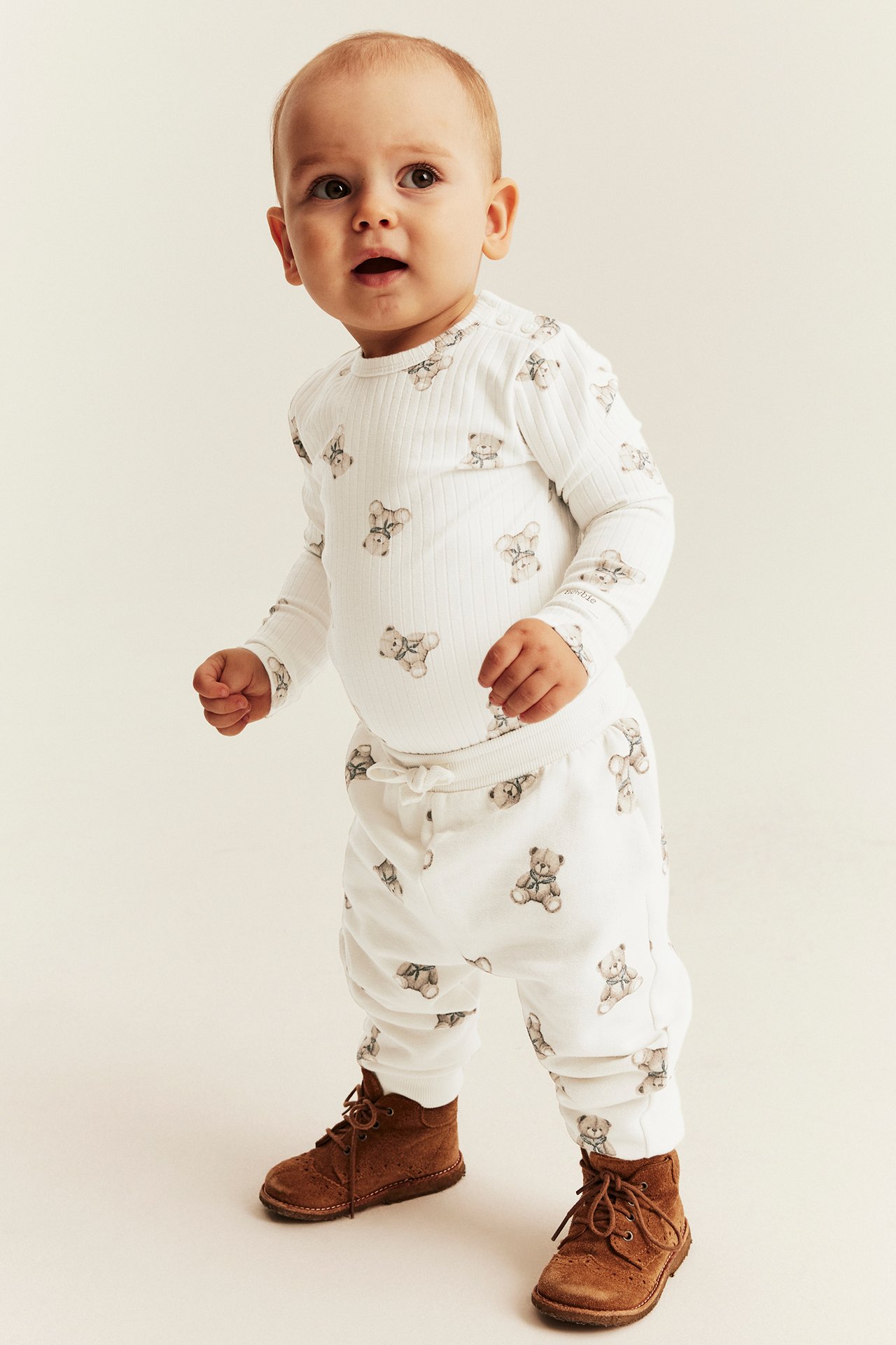 Front view of baby in white ribbed teddy bear print long sleeve top, matching joggers, brown lace-up boots.