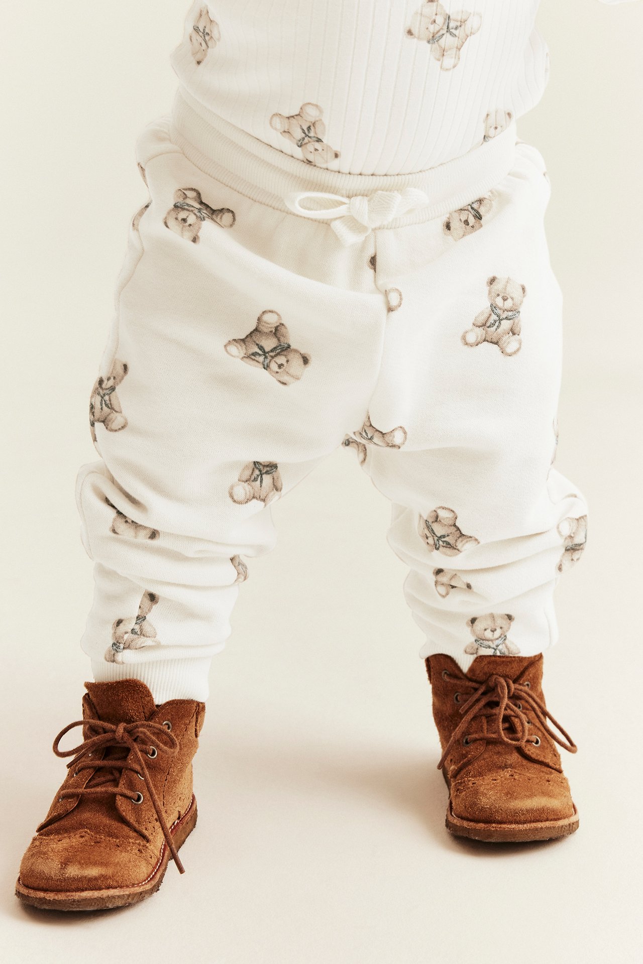 Front view of baby in white teddy bear print sweatpants, matching top, and brown suede lace-up boots.