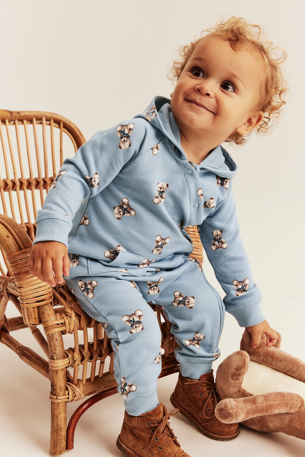 Front view of baby in blue teddy bear print hooded tracksuit, brown boots, holding a beige teddy bear.