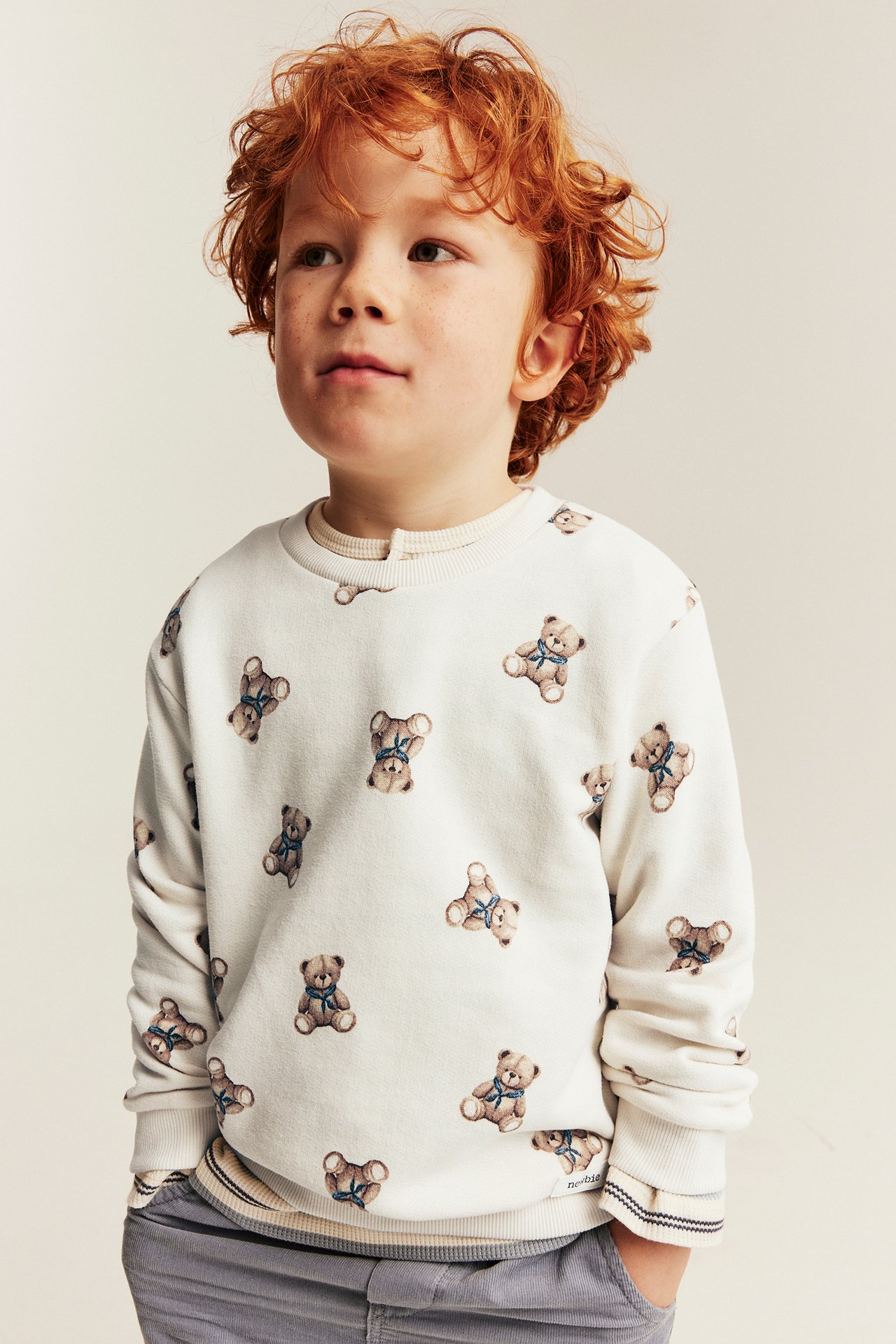 Vorderansicht eines Kindes in weißem Teddybär-Pullover über beigem Oberteil und grauer Hose.