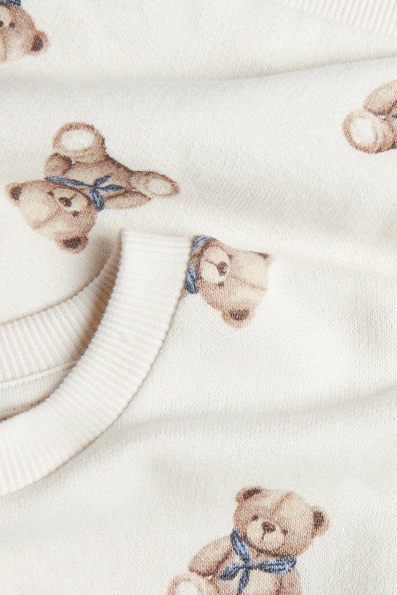 Nahaufnahme eines cremefarbenen Sweatshirts mit charmantem Teddybärenmuster und blauen Schleifen.