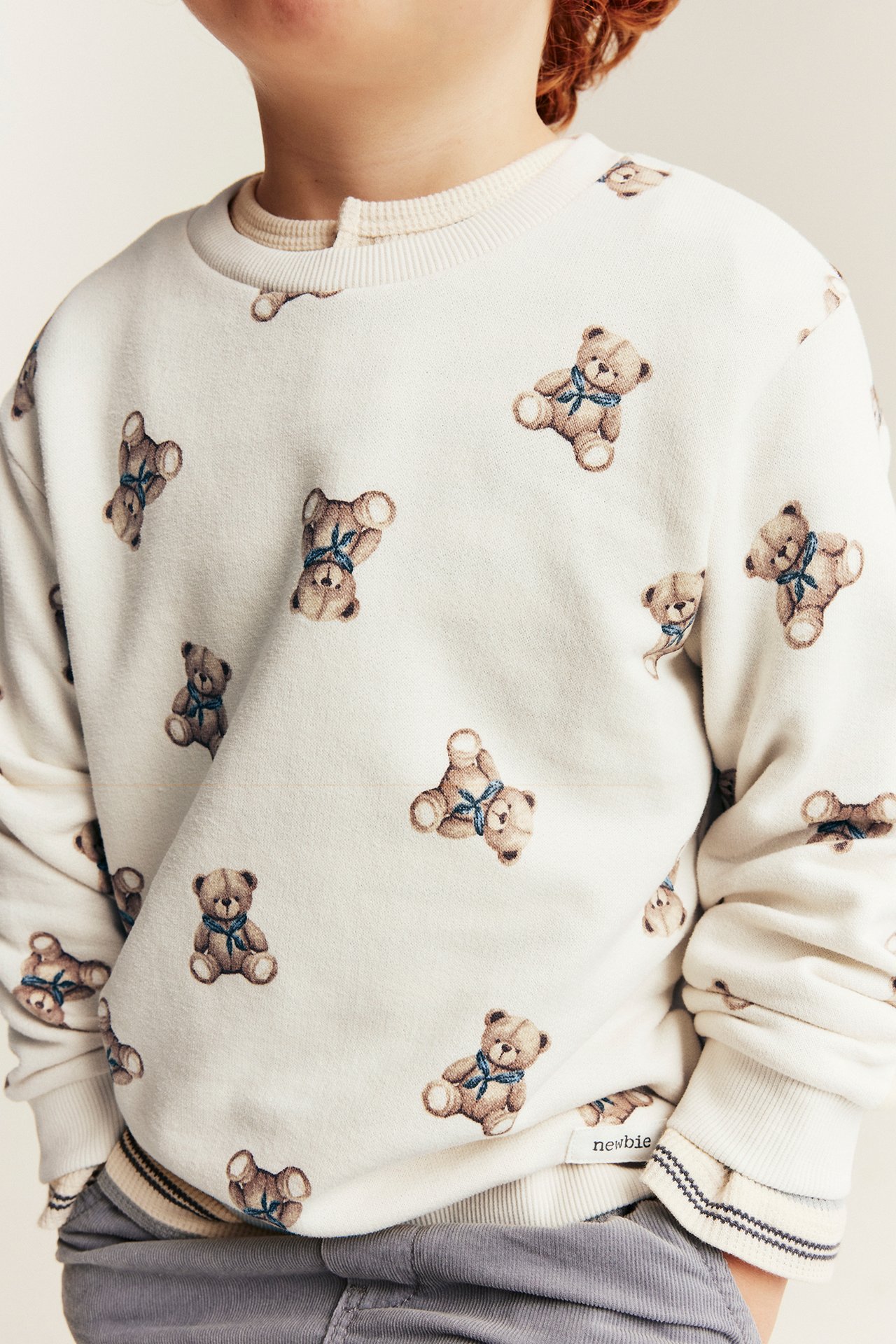 Nahaufnahme eines Kindes in cremefarbenem Teddybär-Print-Sweatshirt, Unterhemd und grauer Cordhose.