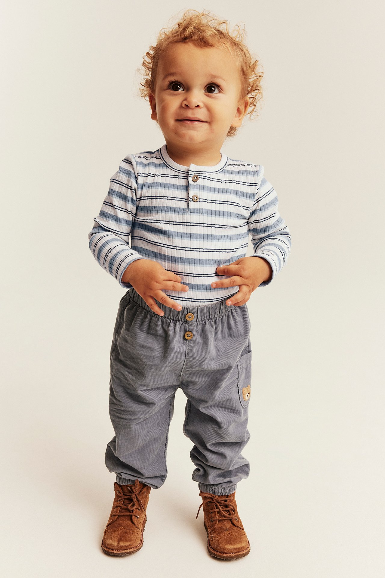 Baby in striped ribbed long-sleeve top, gray elasticated pants with bear patch, and brown boots.