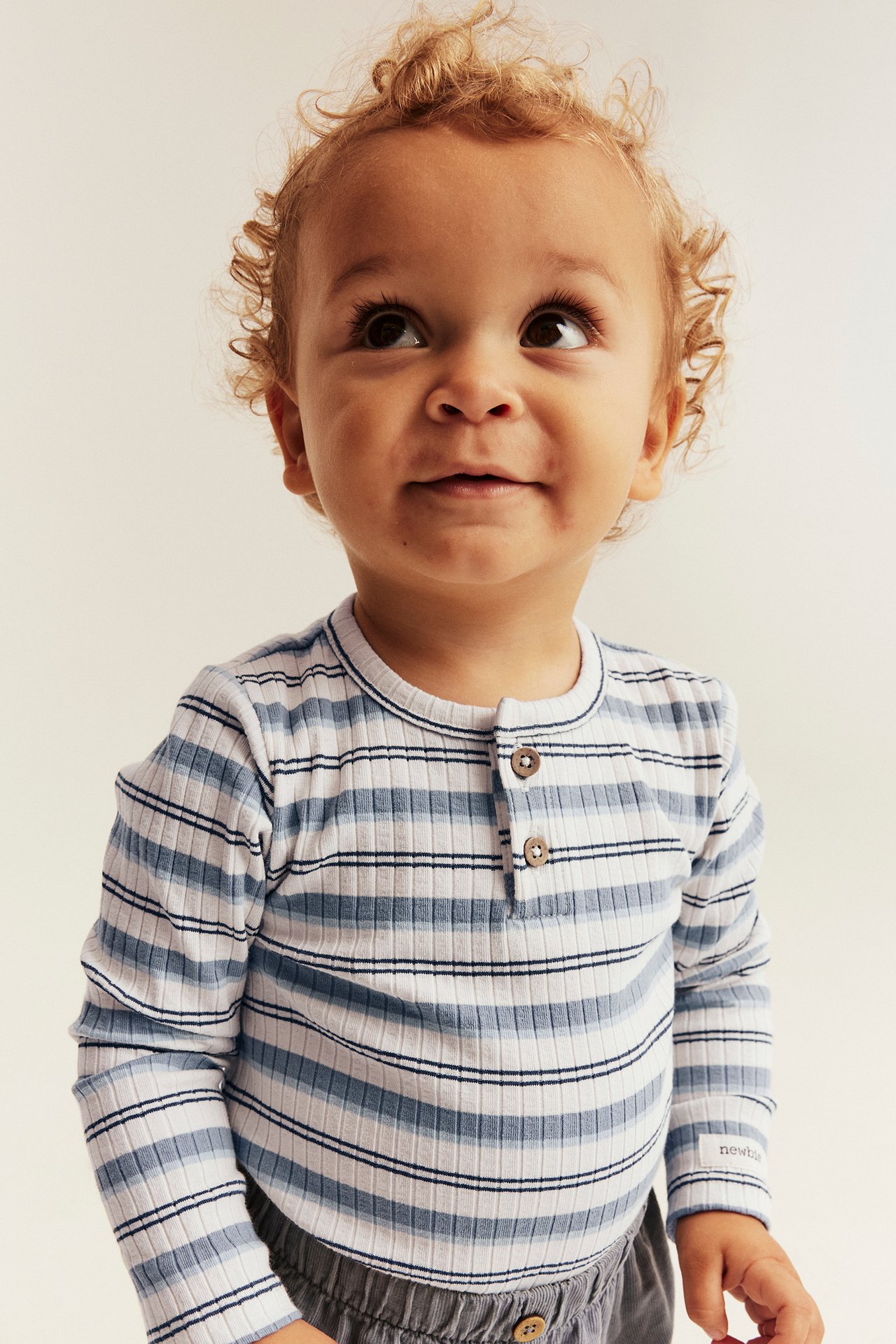 Front view: baby in a blue and white striped ribbed long-sleeved top with buttons and gray pants.