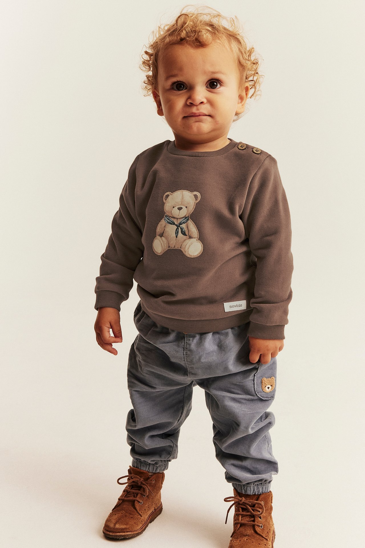 Front view: baby in brown teddy bear sweatshirt, grey corduroy pants, and brown lace-up boots.