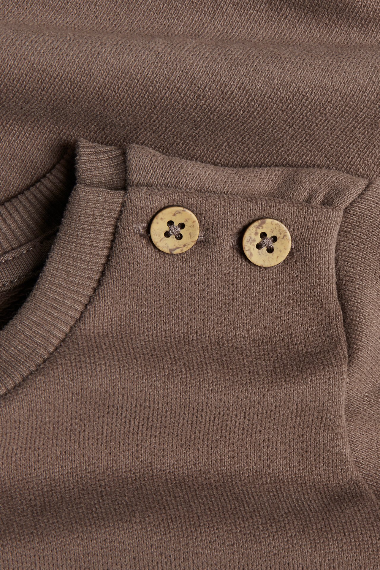 Nahaufnahme eines braunen Strickoberteils mit gerippter Schulter und zwei hellbraunen Knöpfen.