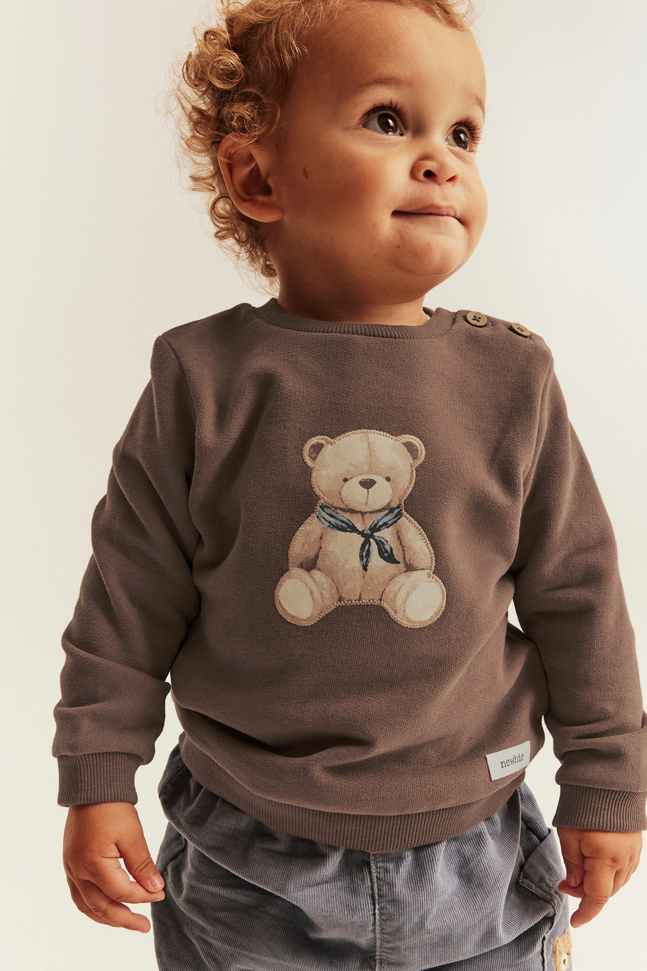 Front view of baby in brown teddy bear sweatshirt with shoulder buttons and grey corduroy pants.
