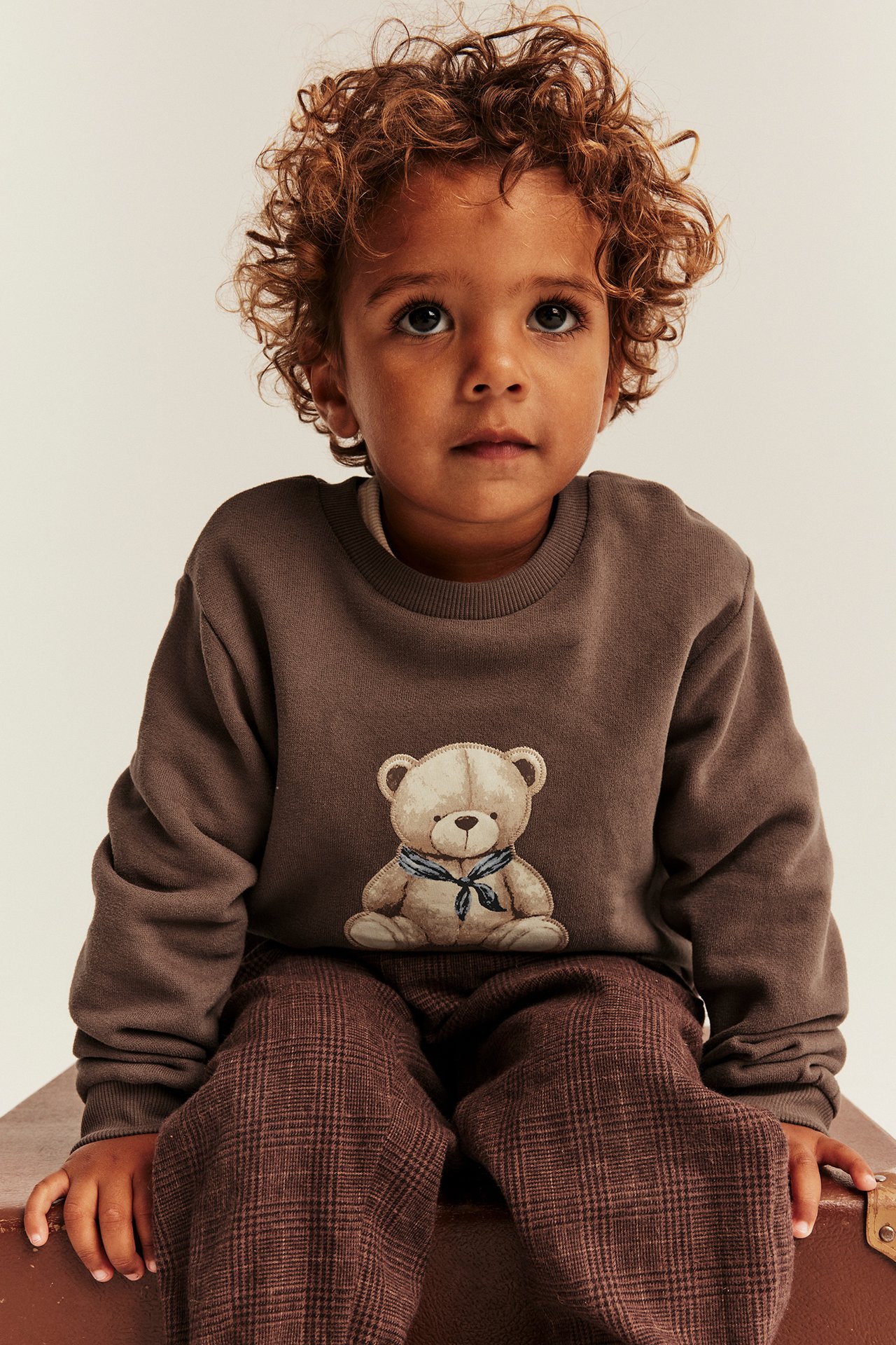 Vorderansicht eines Kindes in braunem Sweatshirt mit Teddybär-Print und brauner Karohose.