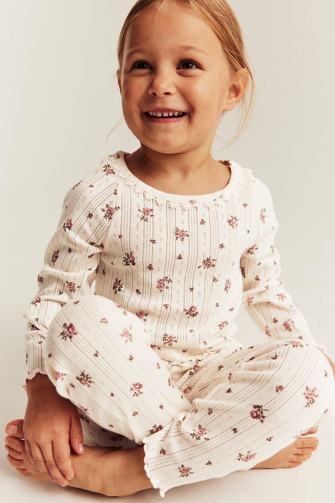 Front view of a kid wearing an off-white floral pointelle knit pyjama set with ruffle trim.